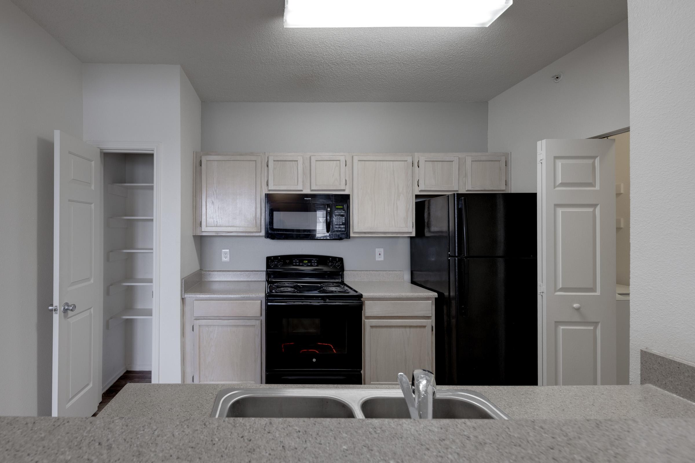 A modern kitchen featuring light-colored wooden cabinets, a black refrigerator, and a black stove. The countertop is made of light stone, and there is a double sink. A pantry and another door leading to a room can be seen in the background. The walls are painted white, and the lighting is bright.