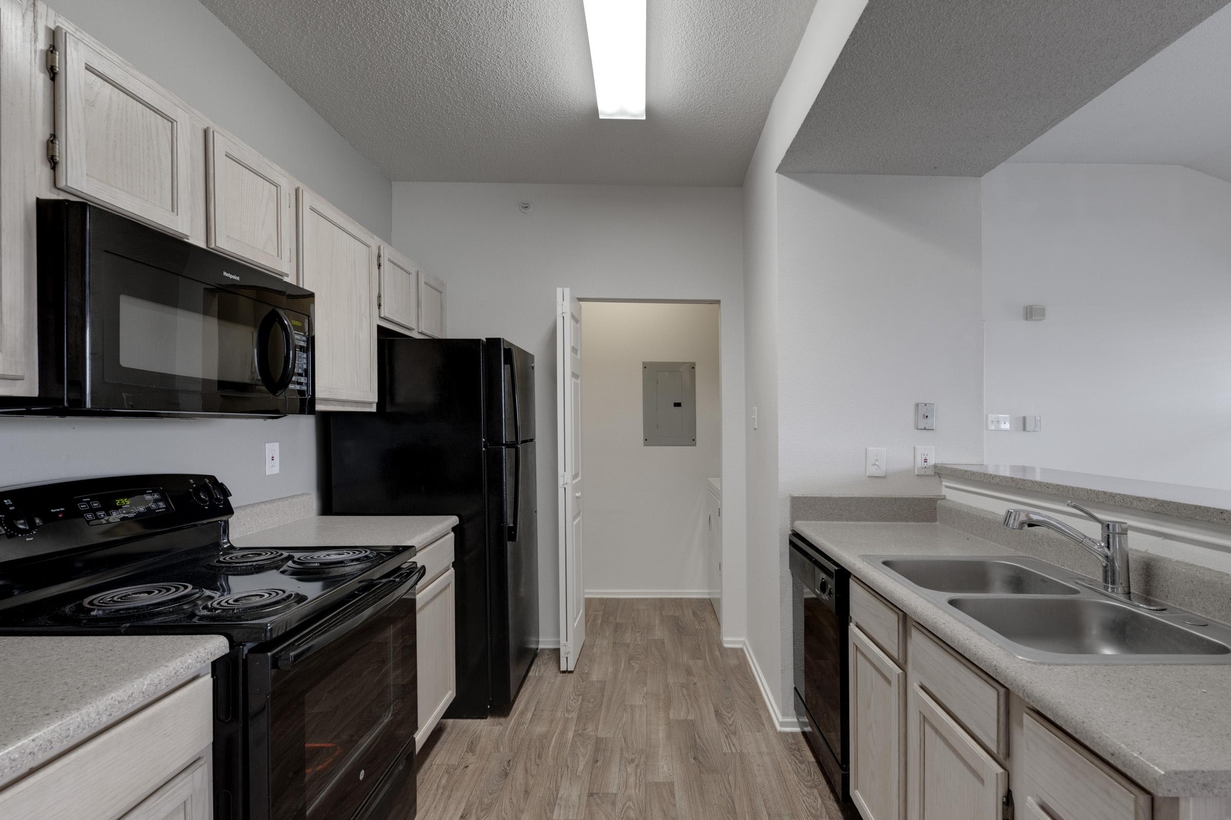 Modern kitchen showcasing light-colored cabinets, a black microwave, and a stove, alongside a black refrigerator. The countertops are beige, with a double sink on the right side. A door leads to another room, and the flooring is a light wood finish. Bright overhead lighting enhances the space.