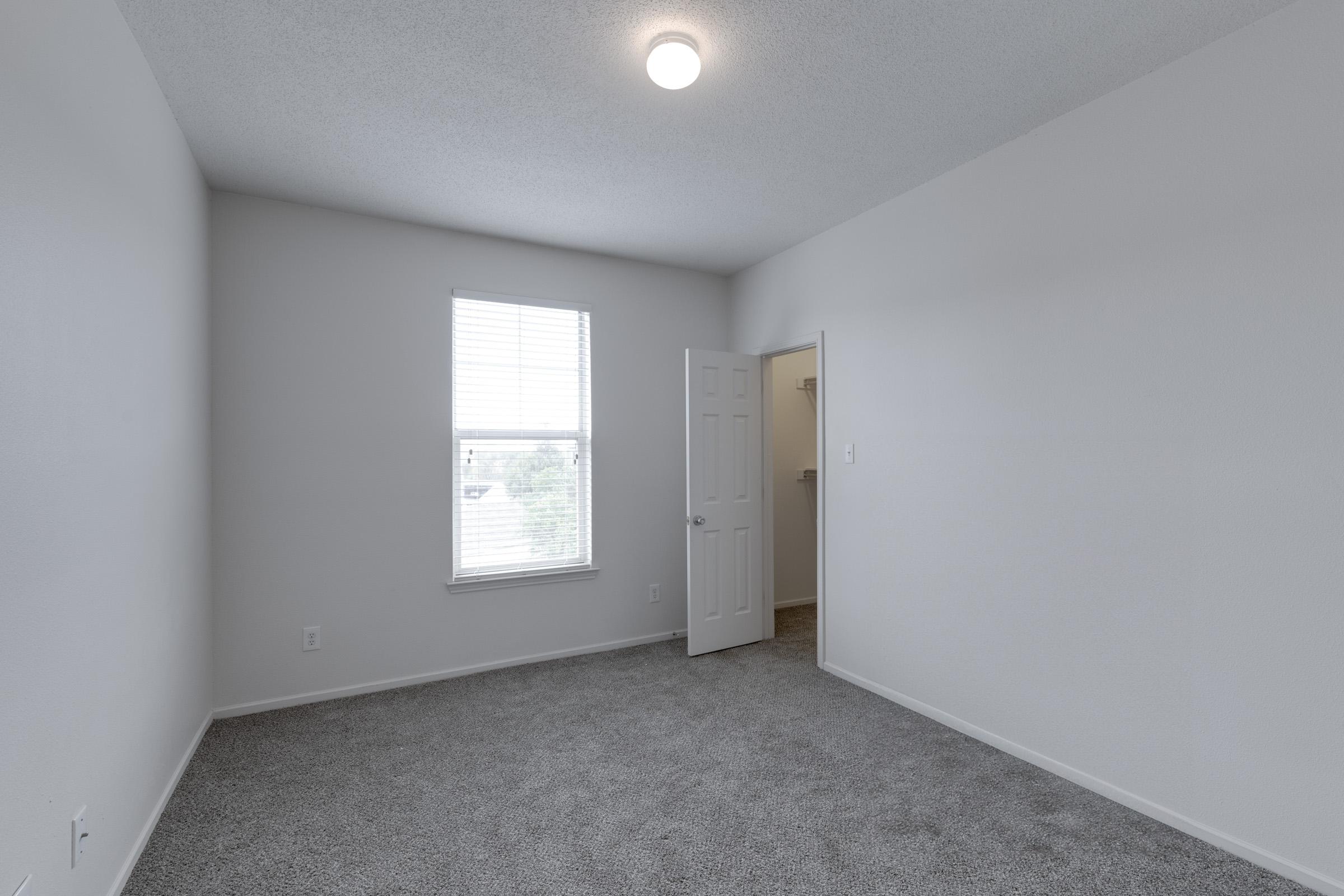 Empty room with light gray carpet, a small window with blinds allowing natural light, a white ceiling with a light fixture, and a door leading to a closet or another space. The walls are painted in a neutral color, creating a simple and spacious atmosphere.