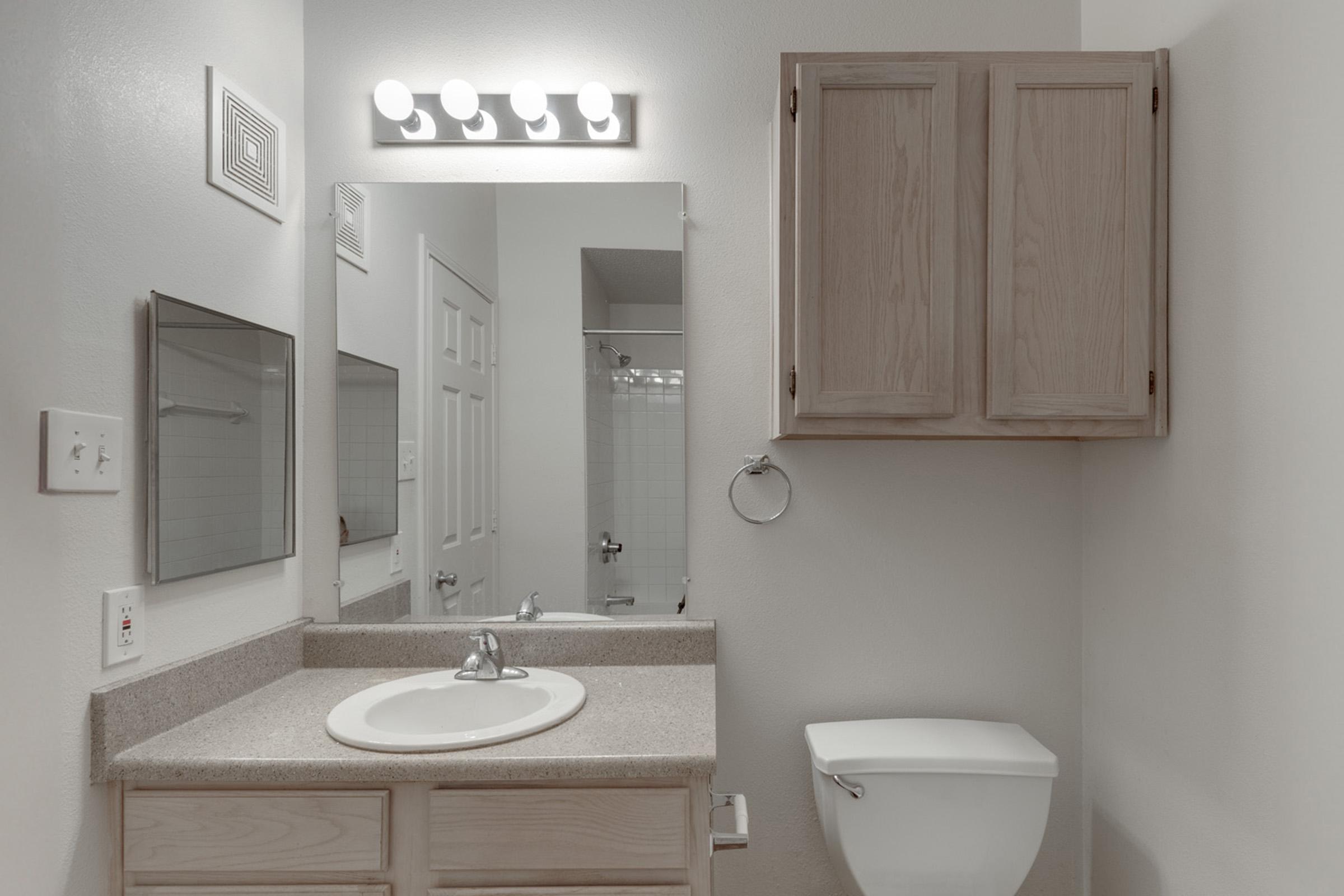 A clean and minimalistic bathroom featuring a countertop with a sink, a mirror with overhead lighting, a wooden cabinet, a toilet, and a towel holder. Light-colored walls and tiles create a bright and spacious atmosphere. A glass shower can be seen in the background.