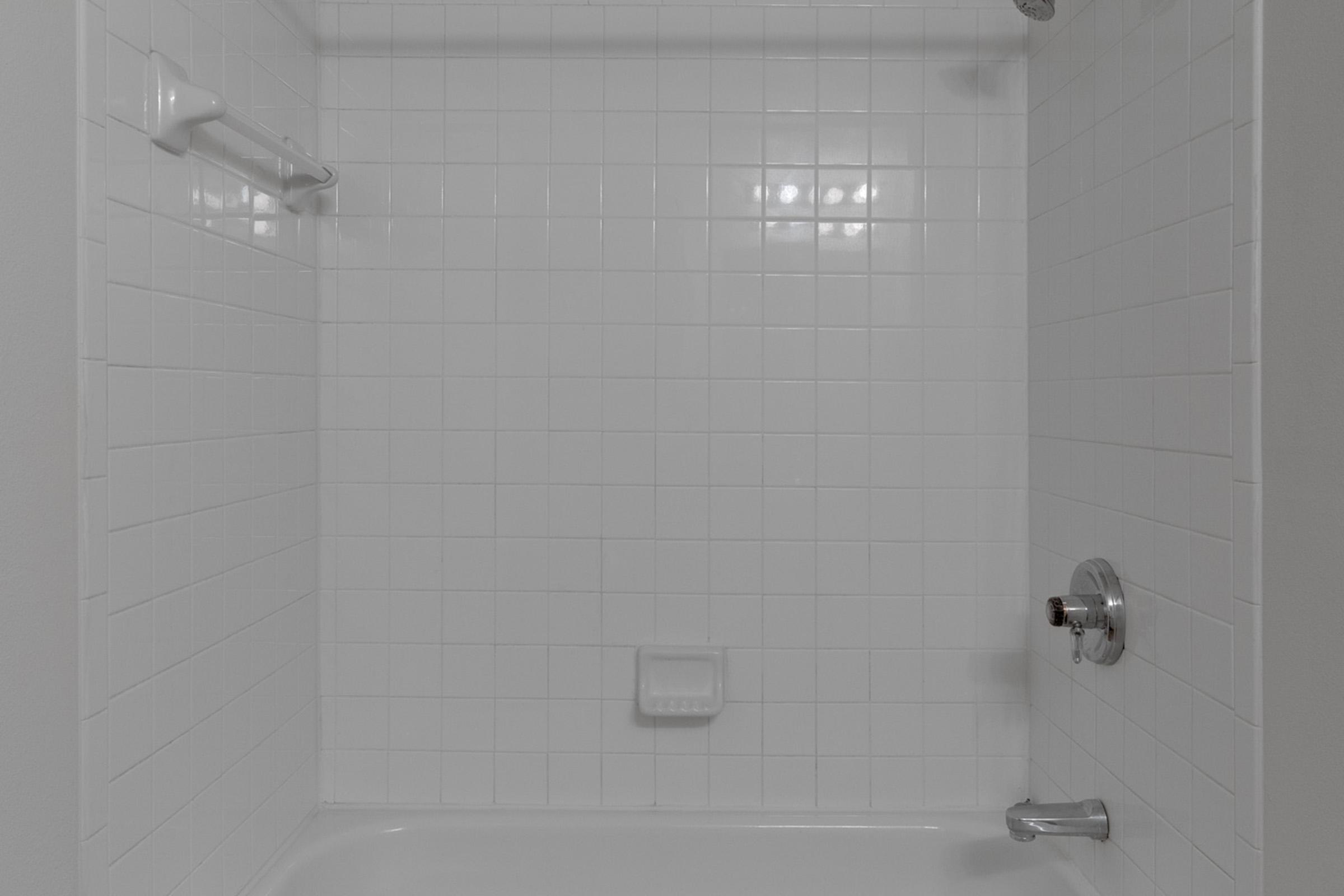 A clean, empty shower stall featuring white tiled walls and a white tub. The showerhead is on the right side, and there's a soap dish on the wall. A towel bar is mounted on the opposite wall, with bright lighting enhancing the simple and minimalist design.