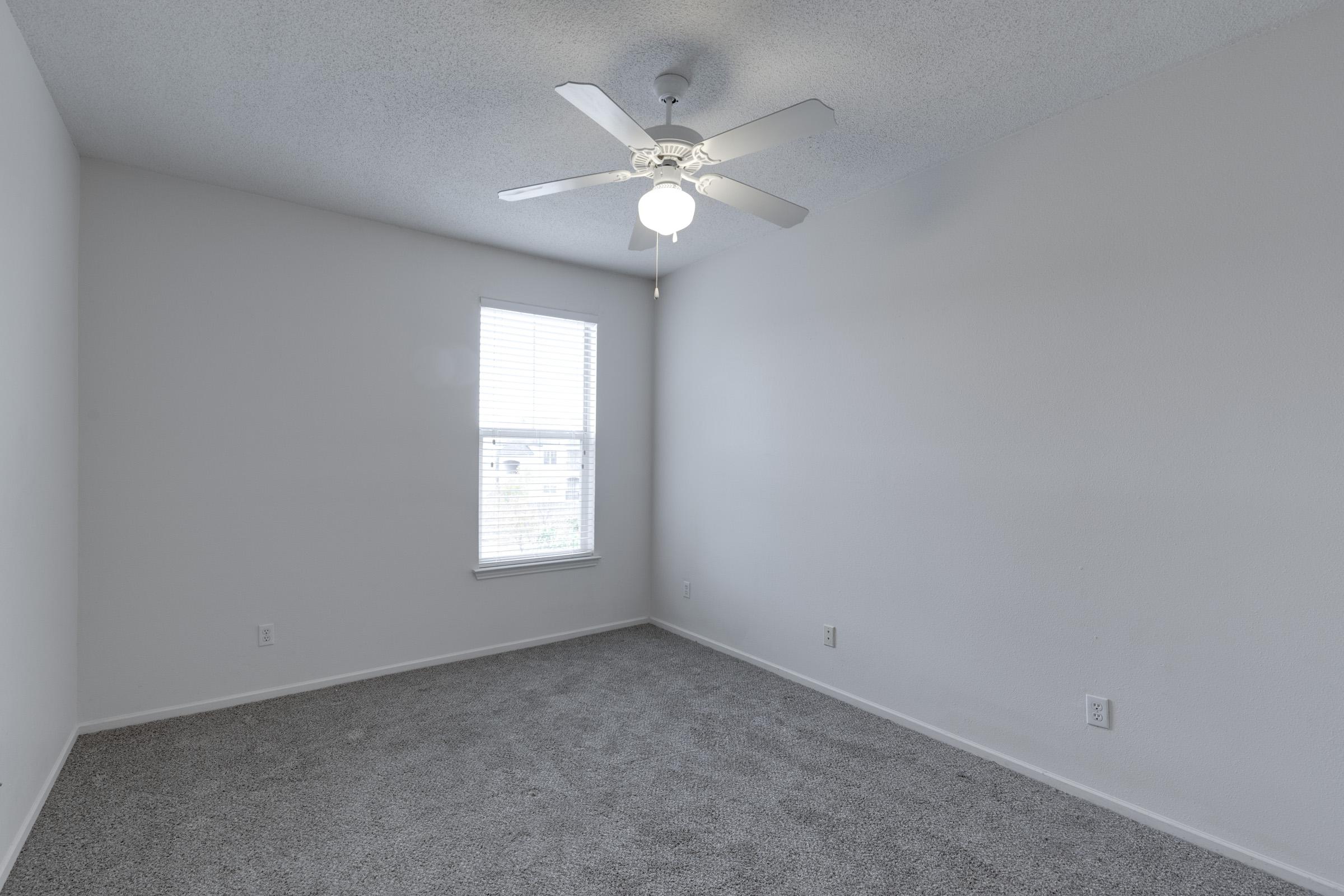 A small, empty room with light gray carpet and plain white walls. A ceiling fan with a light fixture is centered in the room. There is a window on one wall, allowing natural light to enter, and the overall space feels bright and simple, ideal for various uses.