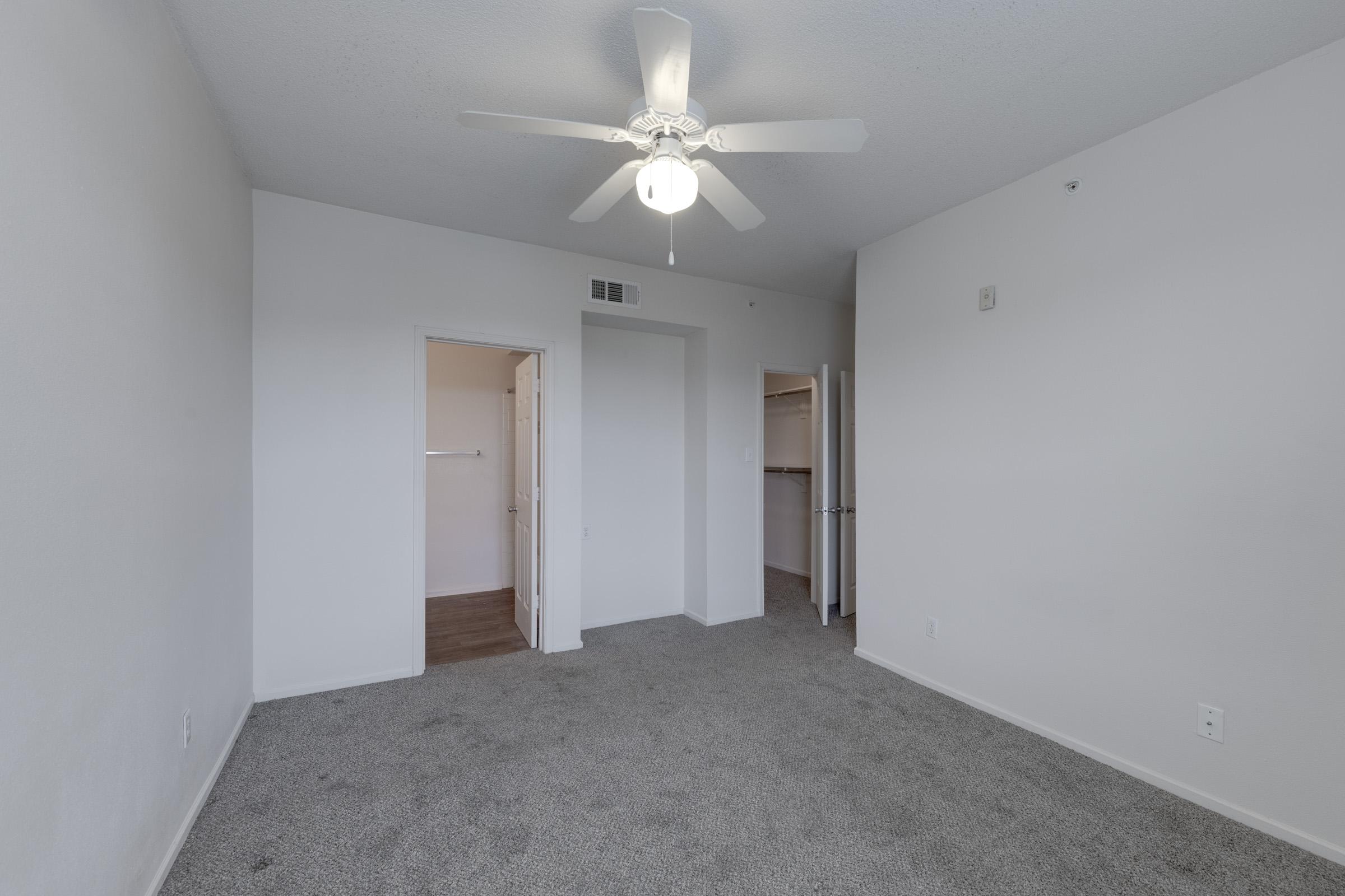 A spacious, empty room featuring light beige carpet, a ceiling fan, and neutral-colored walls. There are two doorways leading to closets or other areas, with one doorway partially visible. The room is well-lit, giving it an open and airy feel.