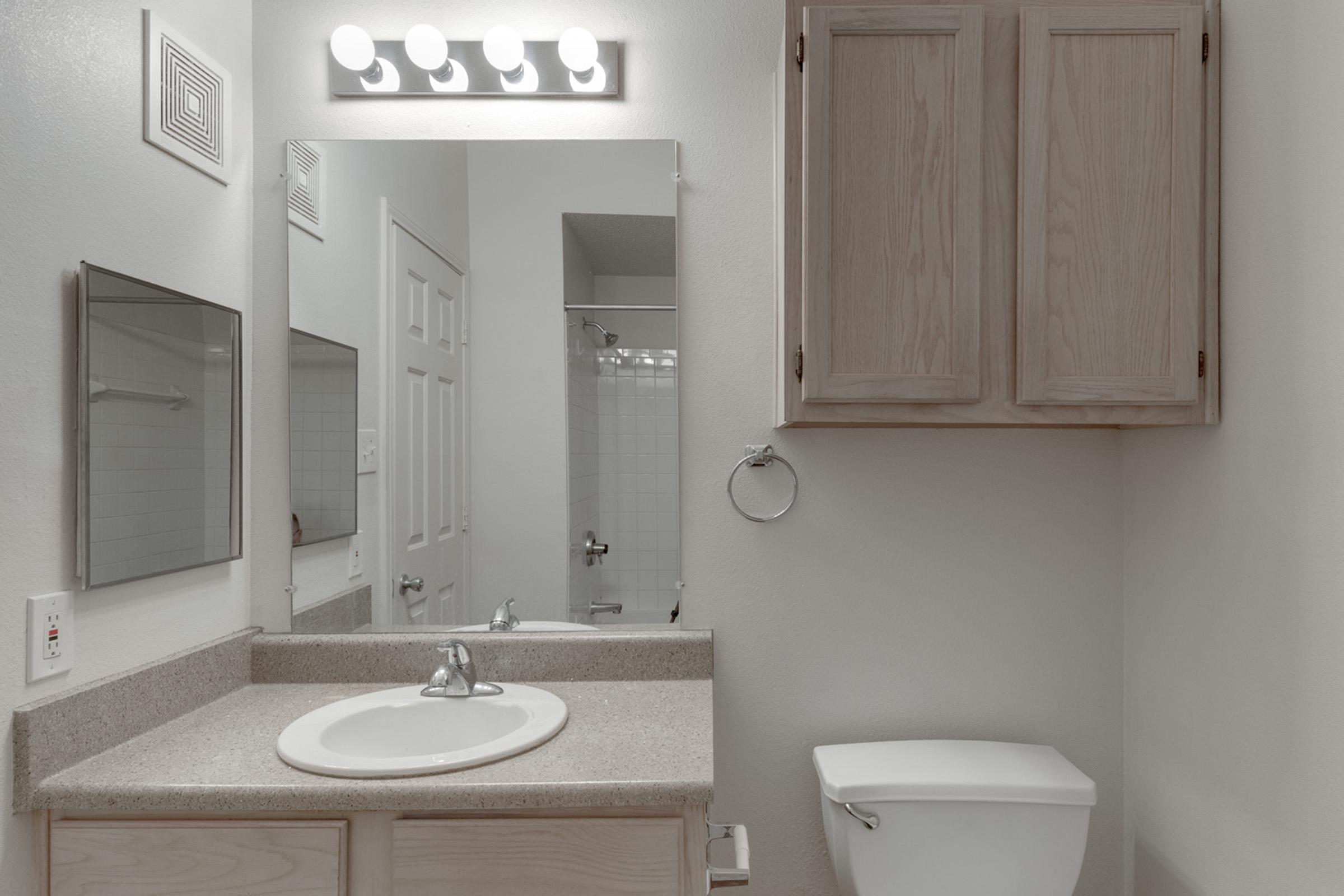 A clean, modern bathroom featuring a countertop with a sink, a wall-mounted mirror, and overhead lighting. A wooden cabinet is mounted above the sink, and a toilet is positioned nearby. The space is well-lit and includes white tiles and a shower area visible in the background.
