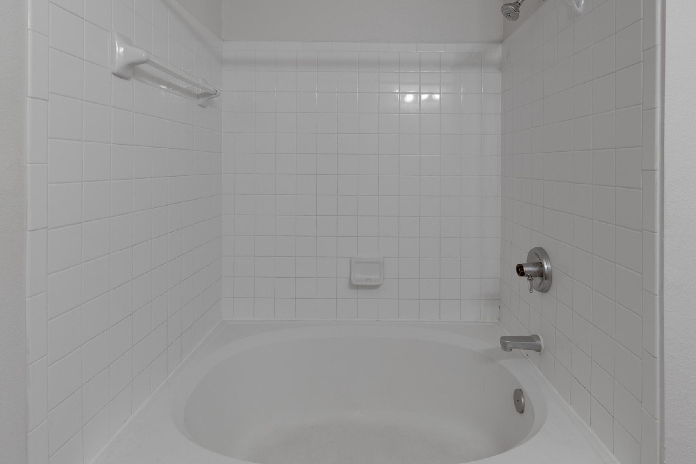 A clean, white bathroom featuring a shower area with tiled walls and a built-in soap dish. The shower has a simple faucet and a grab bar on the wall, with a curved tub bottom, all in a modern, minimalist design. Soft lighting enhances the bright, fresh appearance.