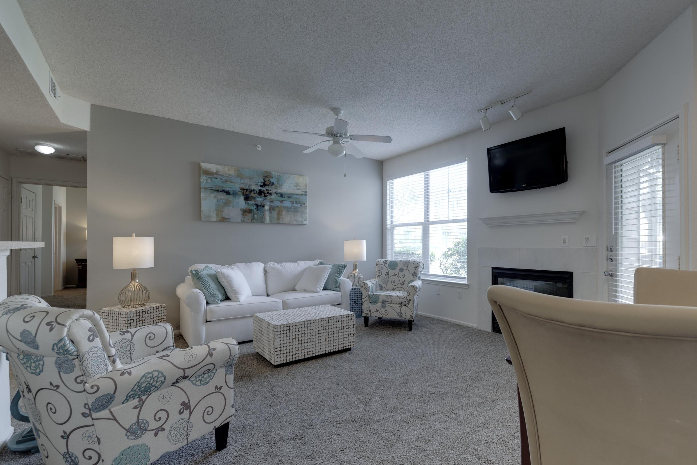 A cozy living room featuring a white sofa with light blue and white accents, two patterned armchairs, and a wicker coffee table. There are large windows allowing natural light, a ceiling fan, and a mounted TV. The space has soft carpeting and neutral walls, creating a warm and inviting atmosphere.