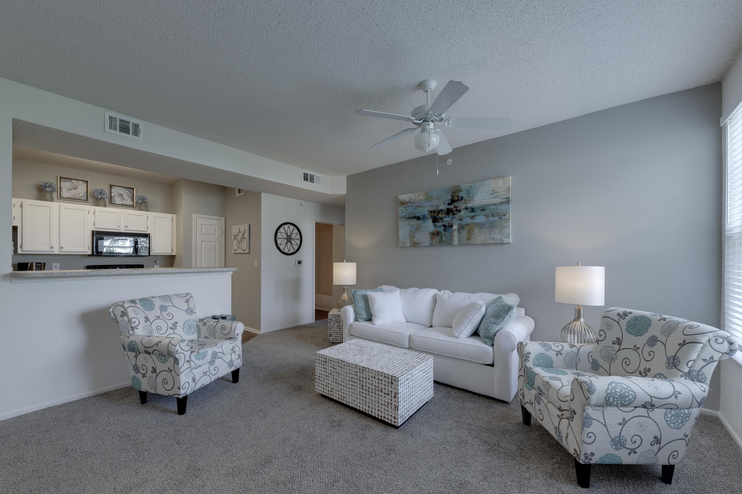Bright and inviting living room featuring a white sofa, two patterned armchairs, and a wicker coffee table. Soft gray walls and carpet create a warm atmosphere, complemented by a ceiling fan and natural light from the windows. A kitchen area is visible in the background, along with a stylish wall clock.