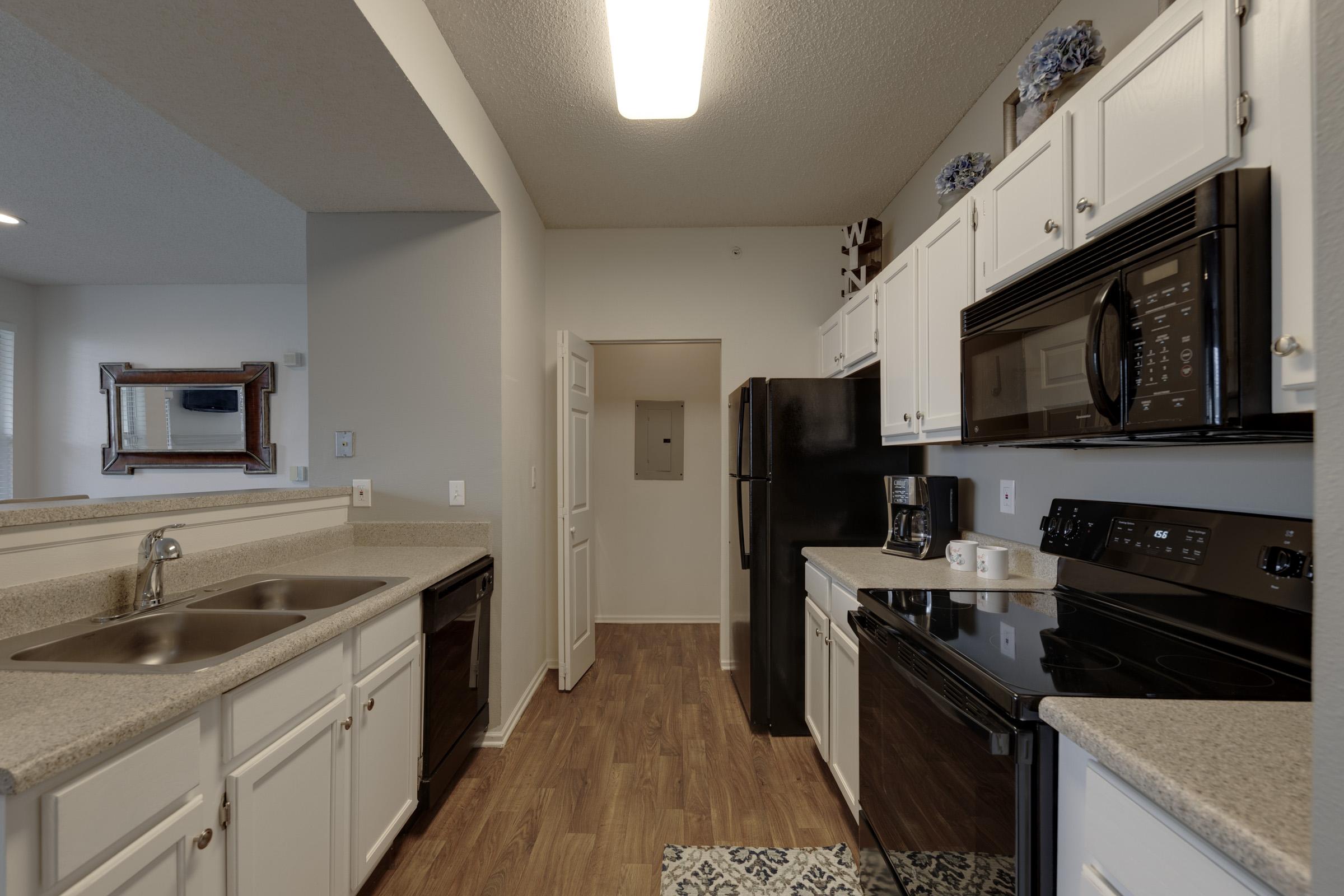 A modern kitchen featuring white cabinets, black appliances, and a countertop. The space includes a double sink, oven, microwave, and refrigerator. Light-colored wood flooring enhances the room, which has a light fixture overhead and is designed with an open layout leading to a hallway.