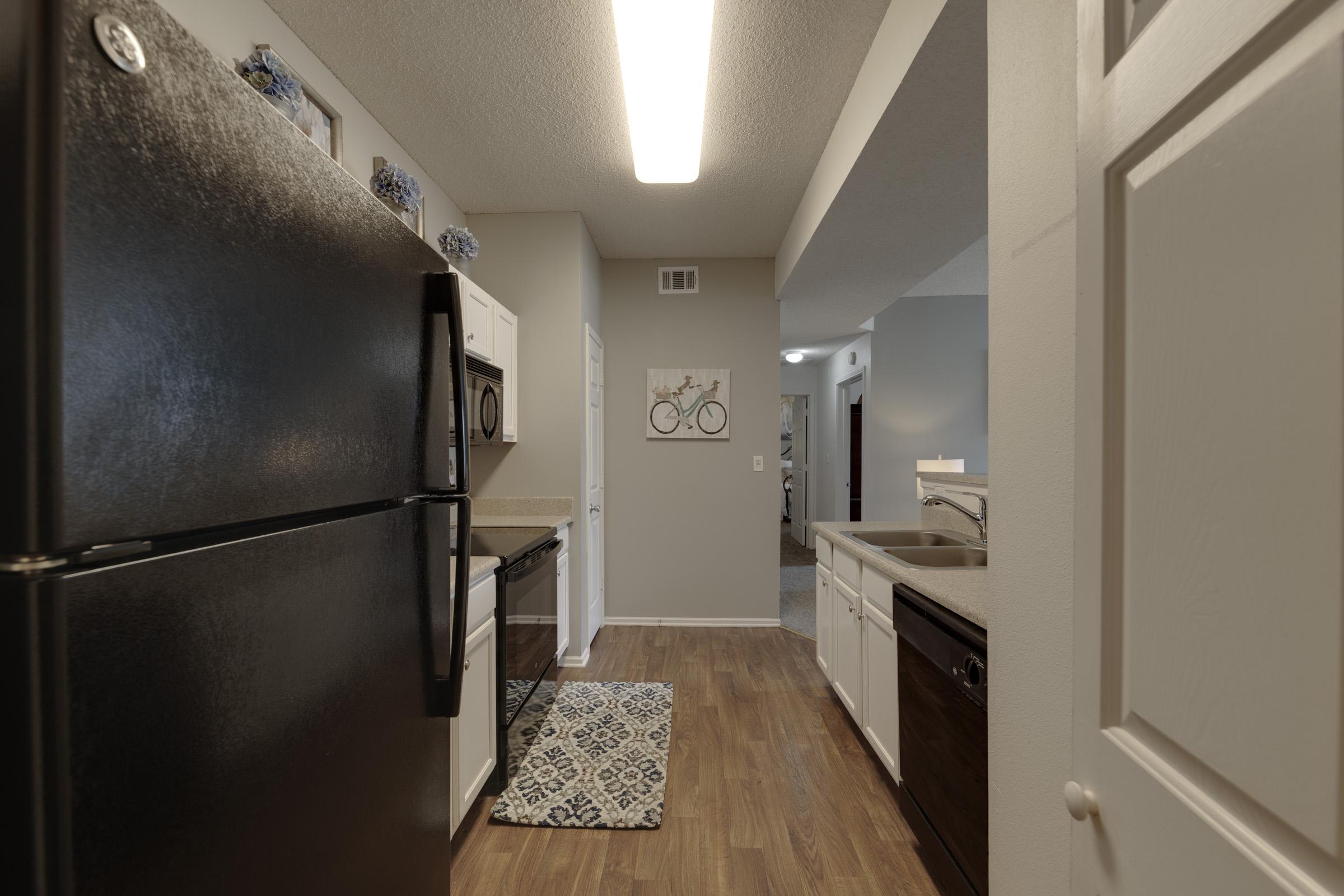 A modern kitchen featuring black appliances, including a refrigerator and microwave, with white cabinets and a light brown countertop. A patterned rug is on the floor, and a bicycle-themed artwork is mounted on the wall. The space is well-lit with overhead lighting and has a neutral color palette.