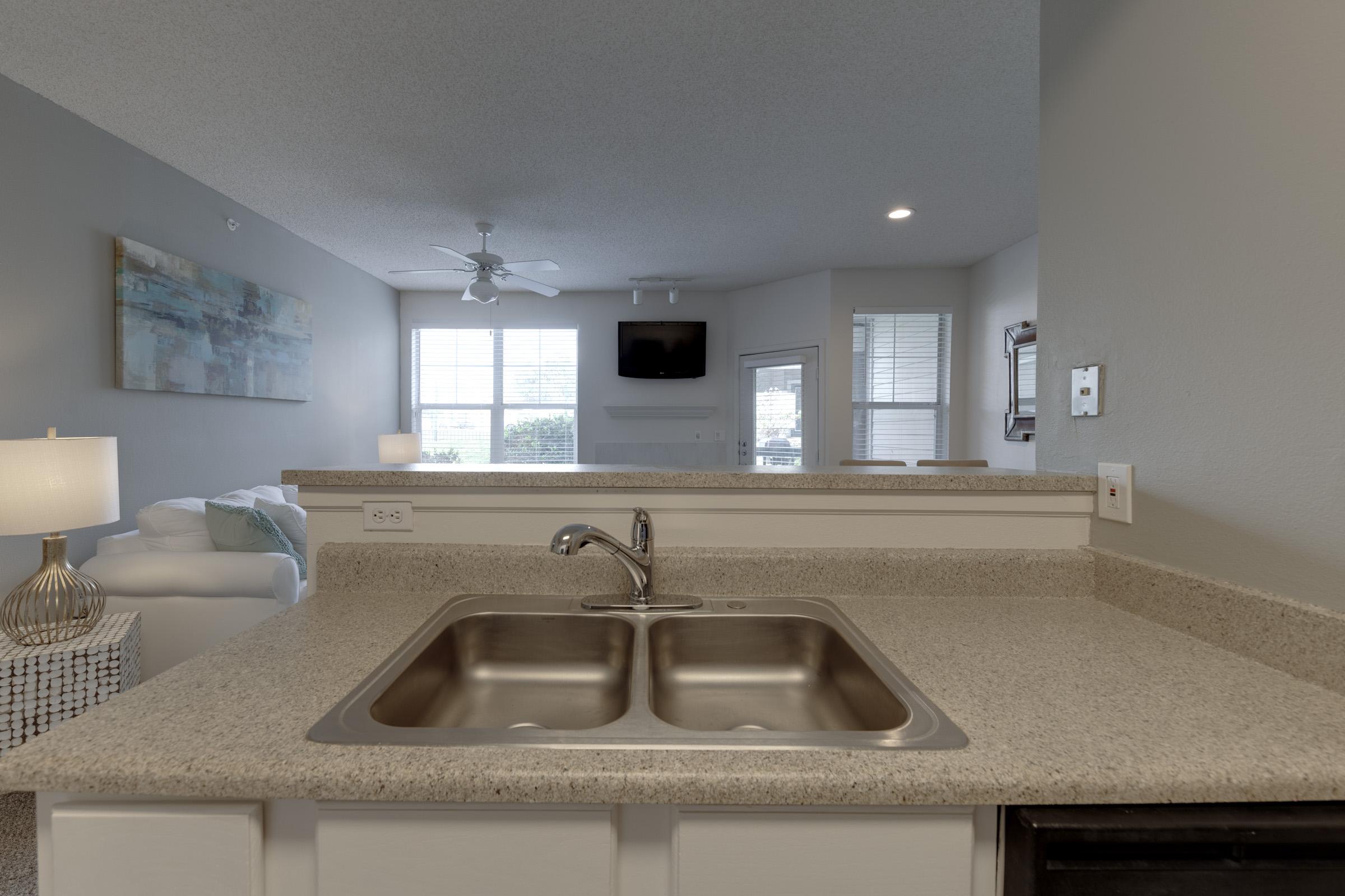 A modern kitchen view featuring a double sink with a stainless steel finish, a light granite countertop, and a cozy living space visible in the background. The living area includes a comfortable white sofa, a ceiling fan, and large windows allowing natural light. Decorative pieces add a touch of style to the space.
