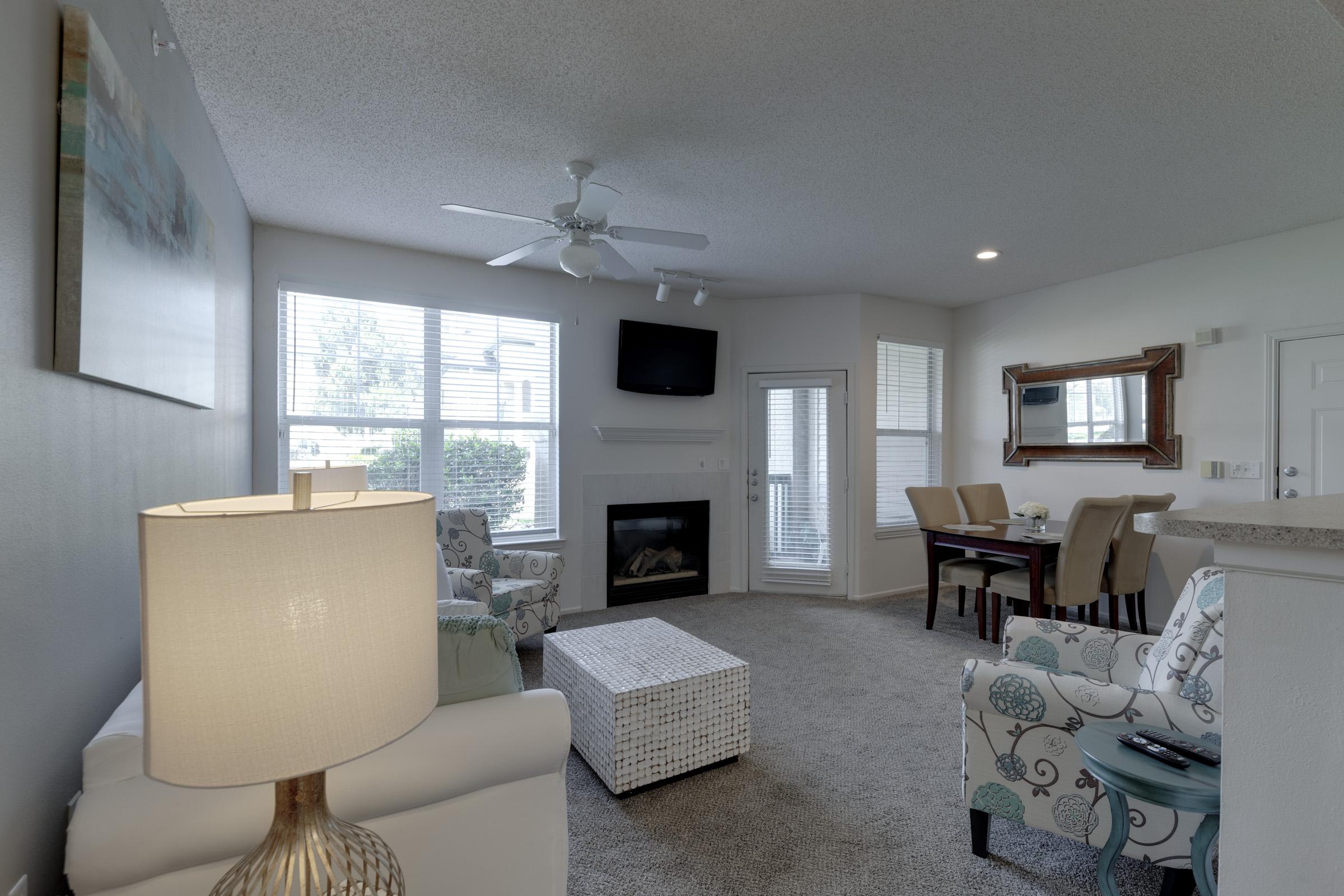 A cozy living room featuring a white sofa, a patterned armchair, and a coffee table. There is a large window with natural light, a ceiling fan, and a fireplace. A dining area with a table and chairs is visible, along with a flat-screen TV mounted on the wall. Soft, neutral décor creates a welcoming atmosphere.