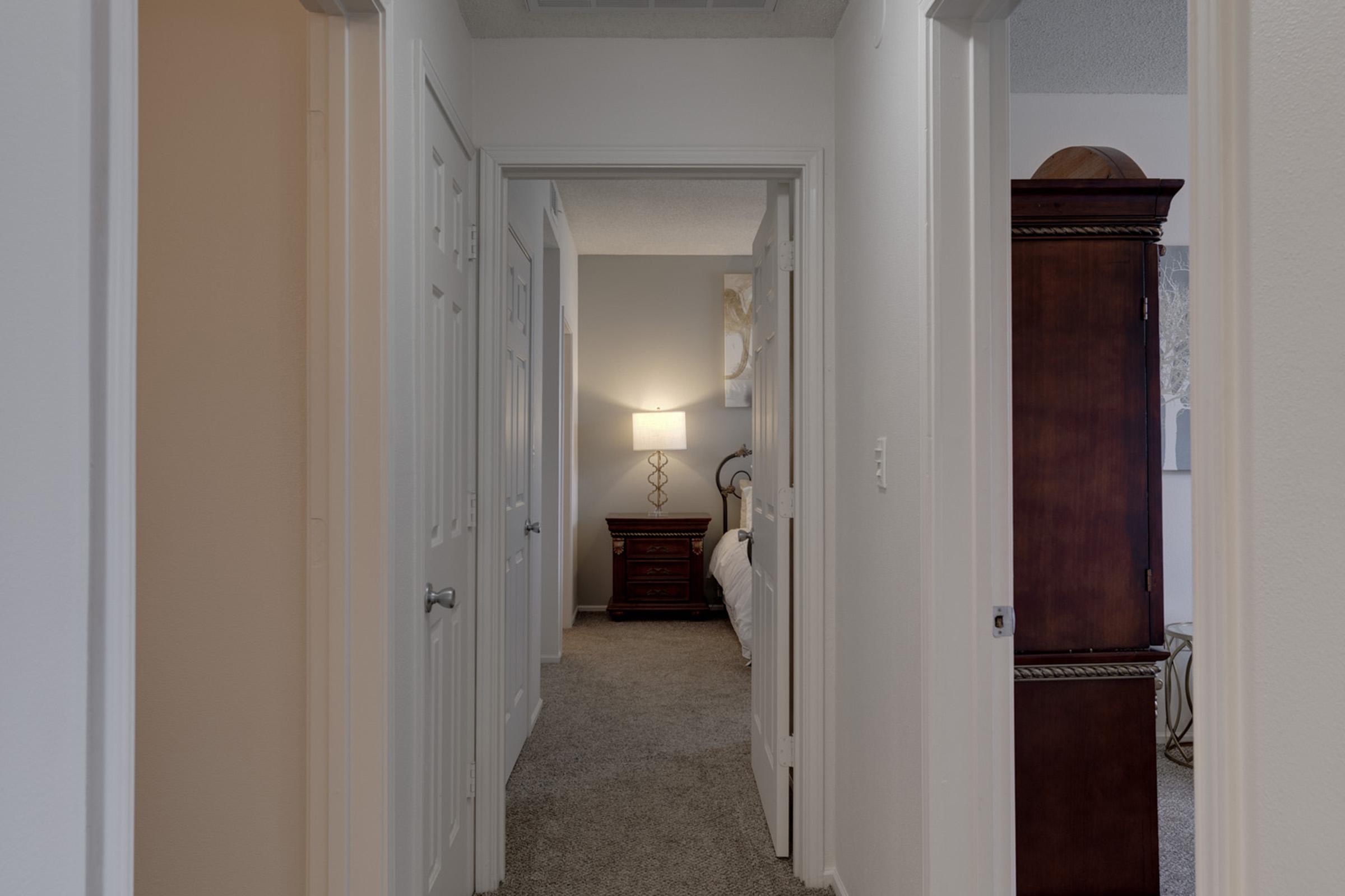 A hallway view with multiple doorways opening to rooms. The walls are painted light colors, and there’s a soft carpet underfoot. At the end of the hall, a lamp illuminates a room, creating a warm ambiance. A wooden dresser is partially visible in one room, adding a touch of decor.