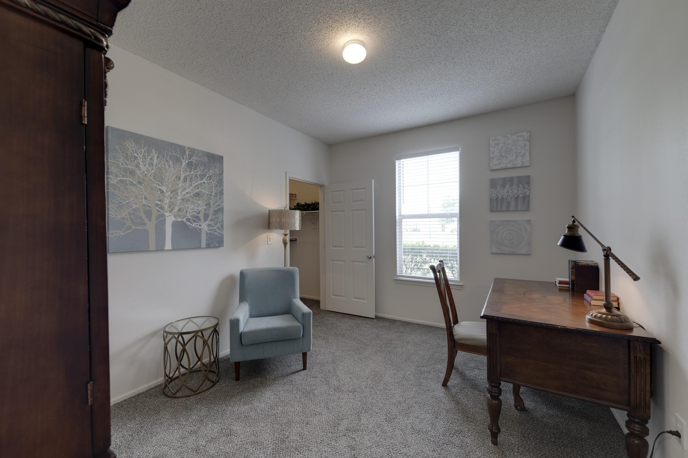 A cozy room with light gray walls and carpet. It features a wooden desk with a lamp, a small blue armchair, a decorative metal side table, and wall art depicting a tree. A door leads to another space, and there's a window allowing natural light in, with a view of greenery outside.