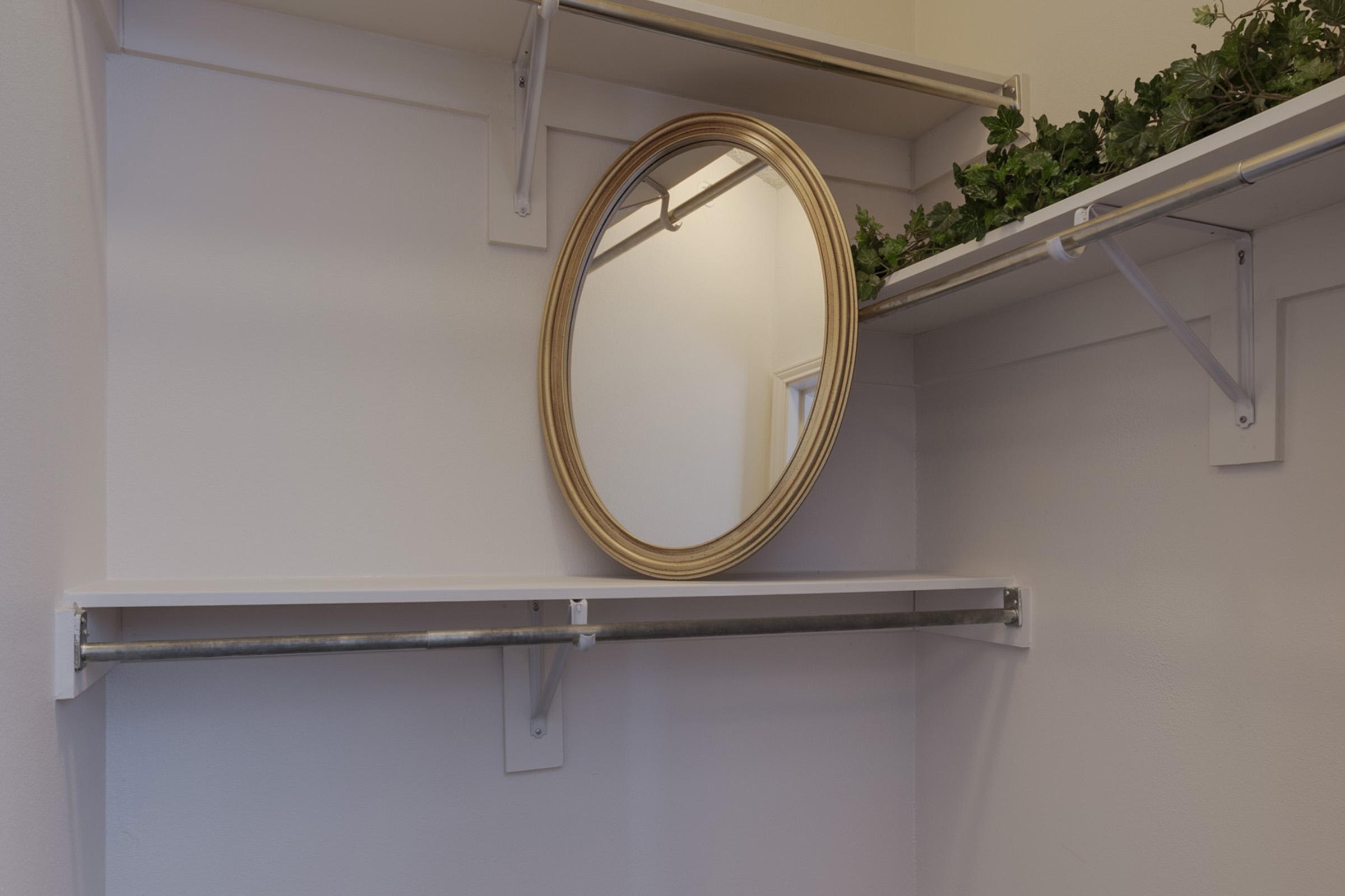 A clean and organized closet space featuring a round gold-framed mirror mounted on a wall. Above the mirror, there’s a small arrangement of green foliage. Two white shelves are visible, providing storage space, while the wall is painted in a neutral color.