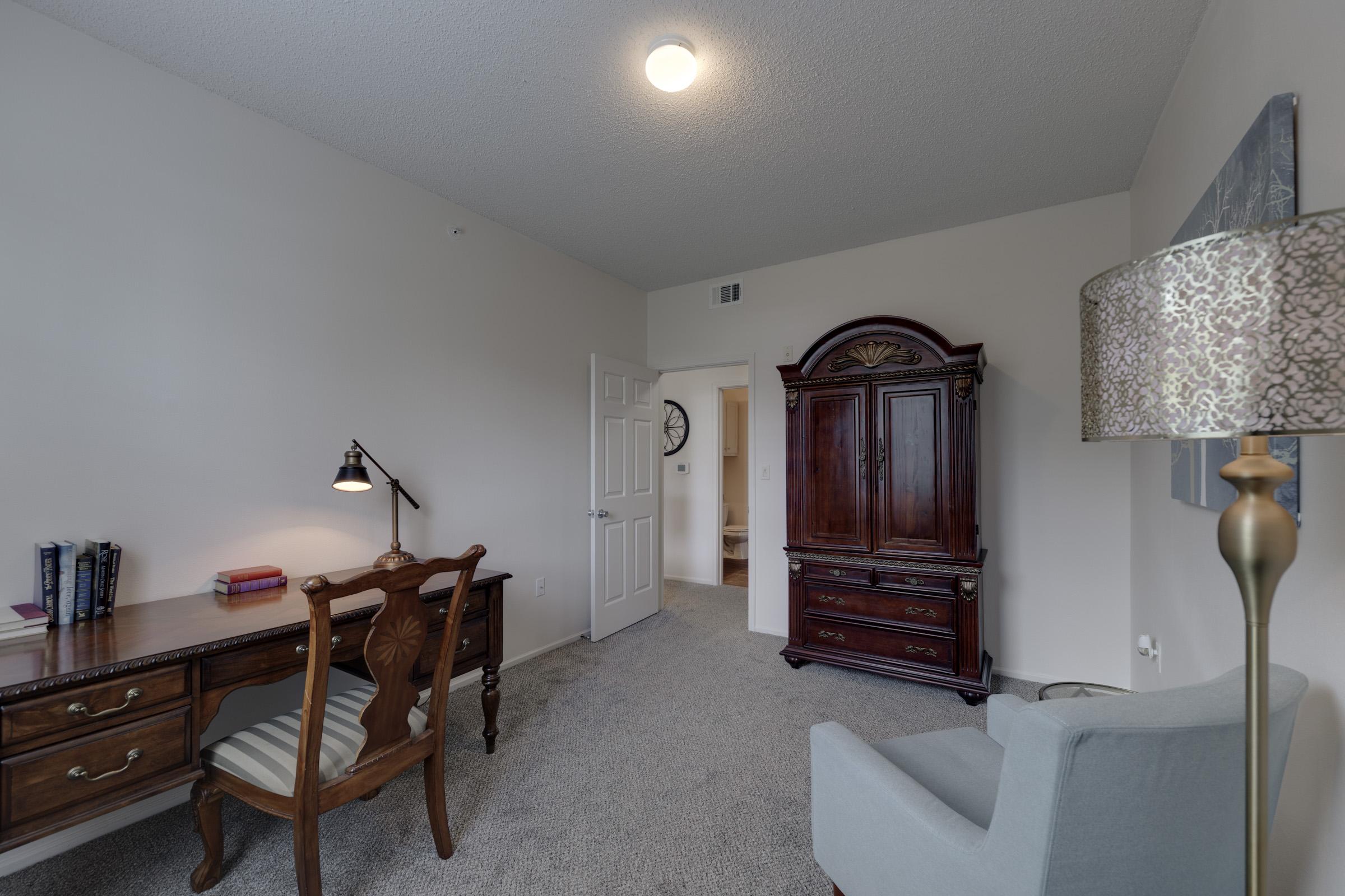 A cozy interior view of a room featuring a wooden desk with a lamp, a comfortable armchair, an ornate dark wood cabinet, and a decorative floor lamp. The walls are painted in neutral tones, and there's a doorway leading to another space in the background. The carpet is light gray, creating a warm atmosphere.