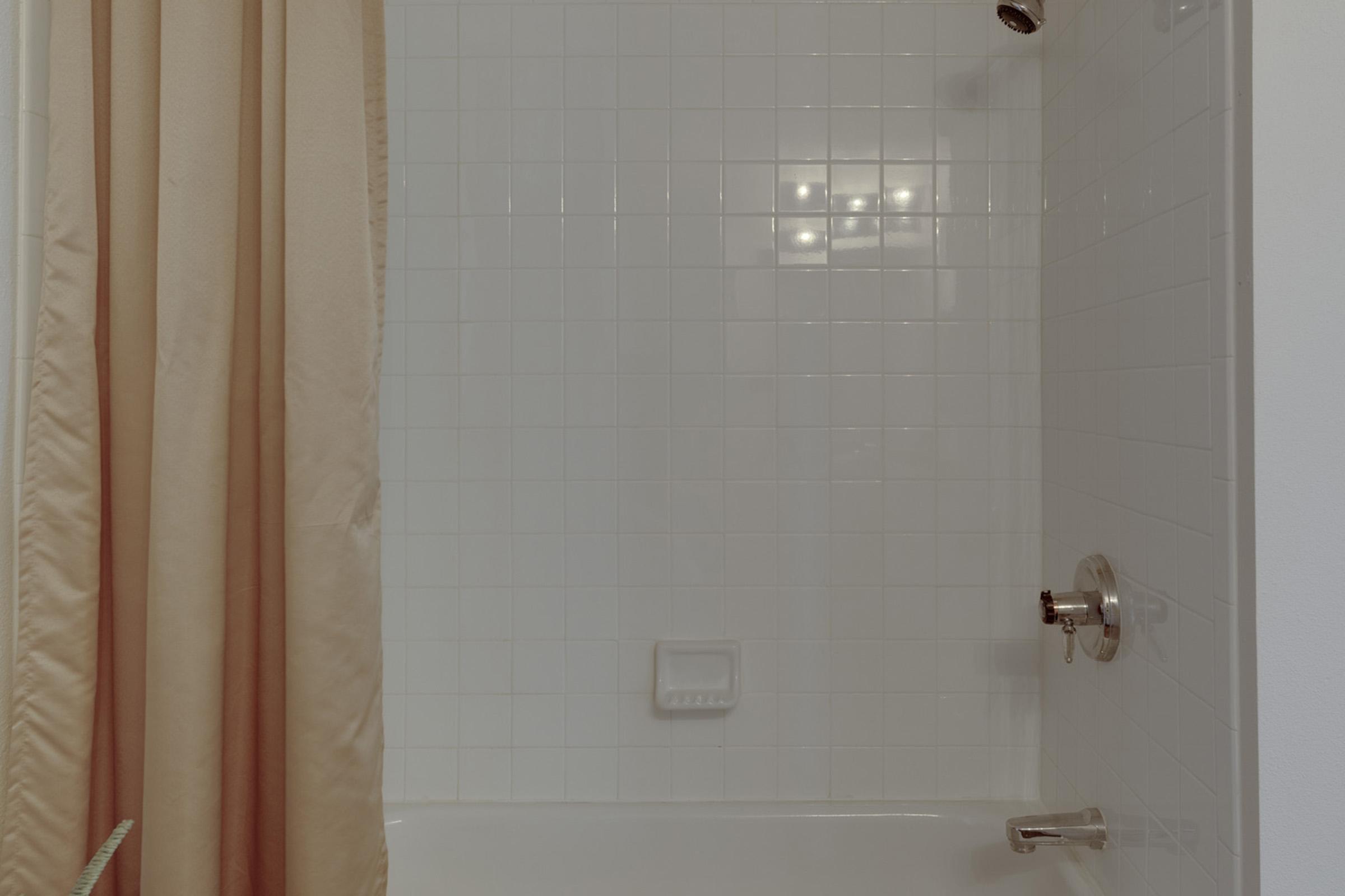 A clean bathroom shower area featuring white tiled walls and a light beige shower curtain. The space includes a silver showerhead and faucet, with a soap dish mounted on the wall. Soft lighting illuminates the area, creating a bright and inviting atmosphere.