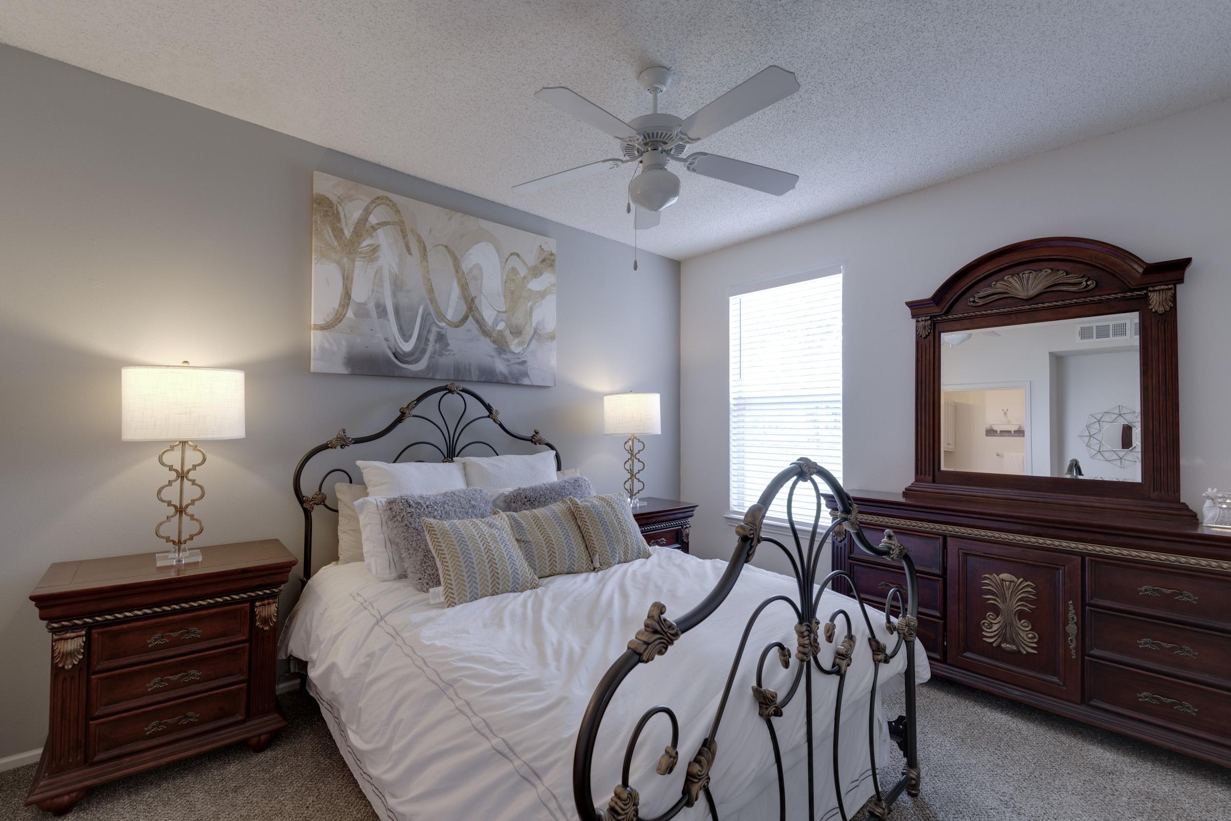 A cozy bedroom featuring a decorative wrought-iron bed frame with white bedding, two elegant nightstands with lamps, a wooden dresser with ornate details, a large mirror, and a ceiling fan. The room is brightened by natural light from a window, complemented by modern artwork on the wall.