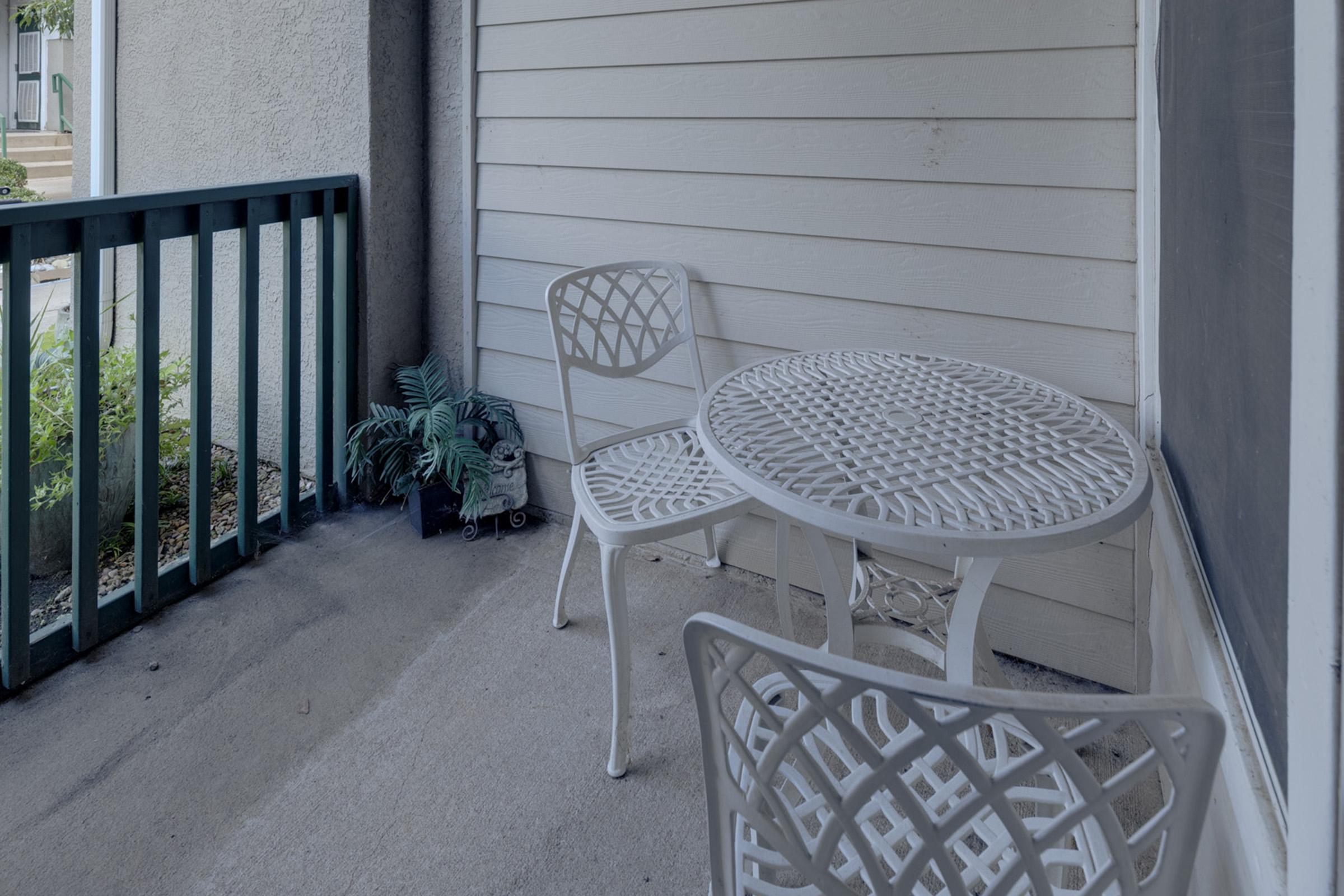A small outdoor patio area featuring a white metal table and two matching chairs. The table has a geometric design on its surface. A potted plant is partially visible in the corner, and there is a wooden railing painted in dark green, contrasting with the light-colored walls.