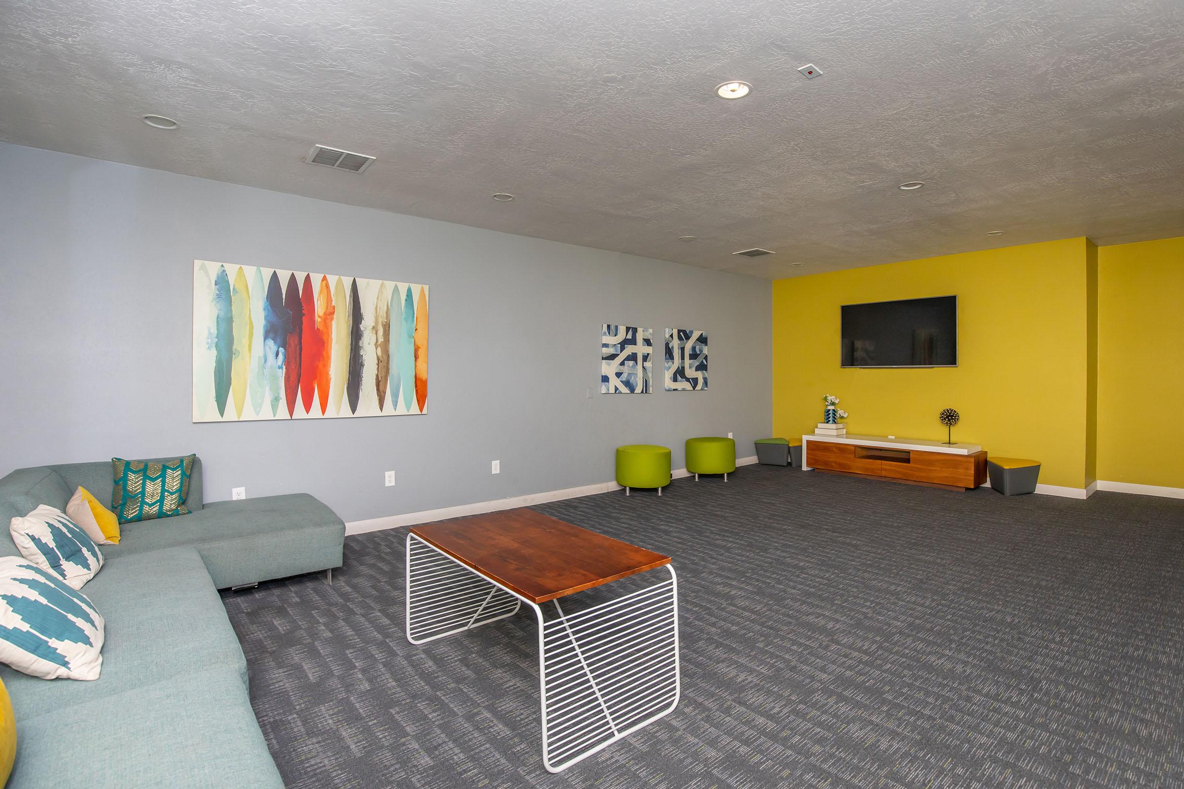Indoor lounge at Rise at the District apartments featuring a sectional couch, coffee table, and flat screen TV on a yellow wall