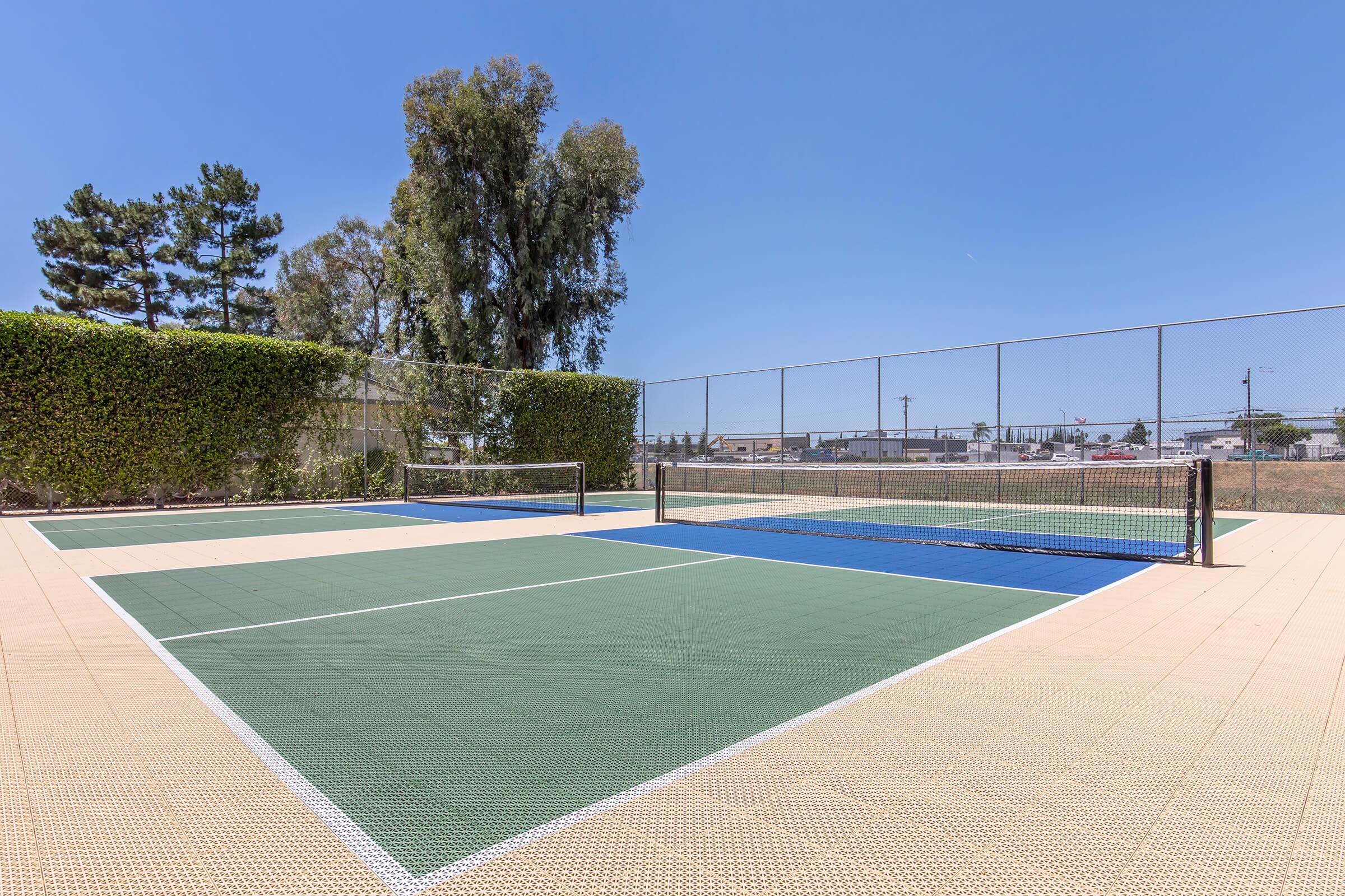 Two tennis courts with green and blue surfaces, surrounded by low shrubs and tall trees. The sky is clear and blue, creating a bright atmosphere. In the background, there's a fence and hints of a nearby industrial area. The courts appear well-maintained and ready for play.