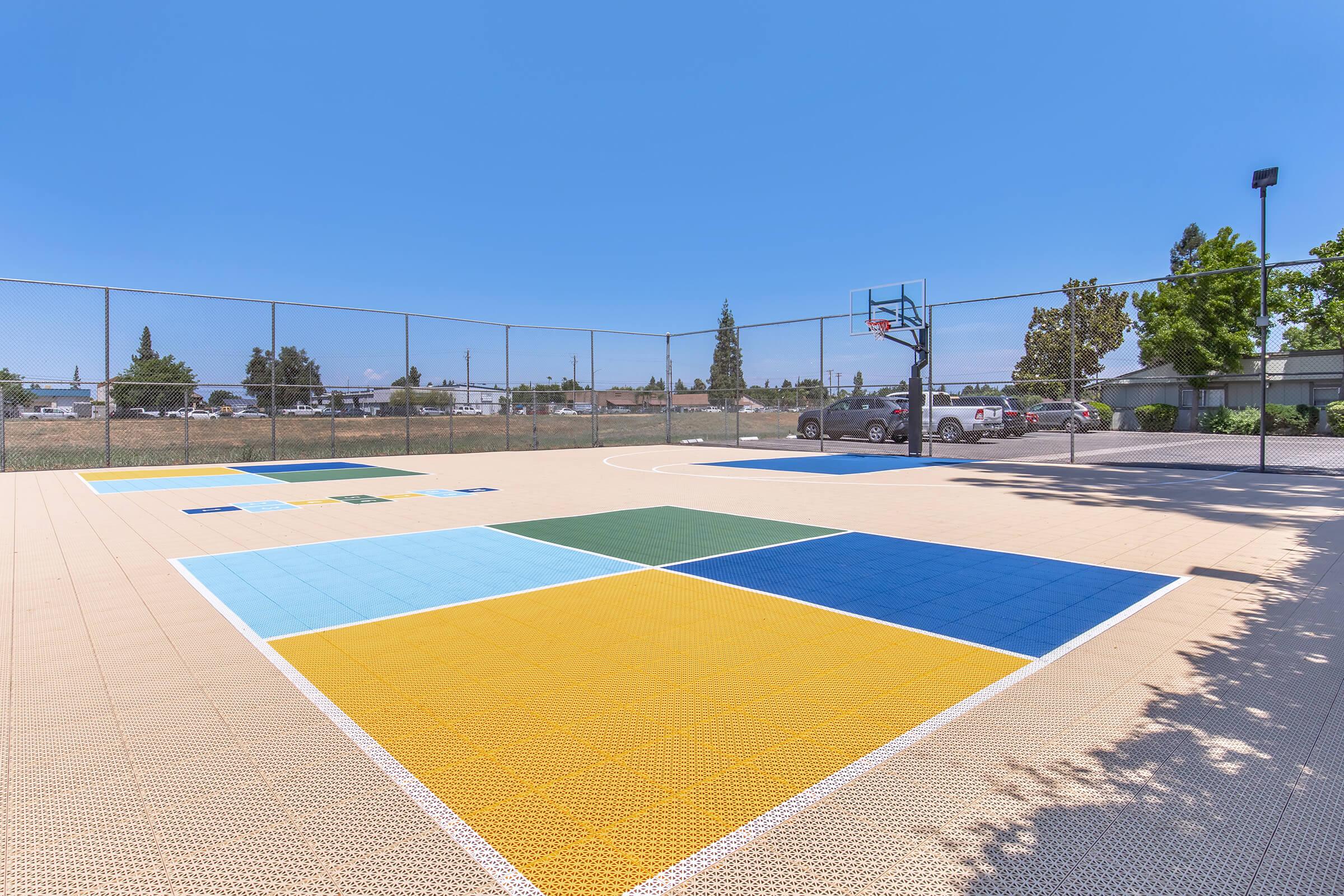 A vibrant outdoor basketball court featuring colored sections in blue, green, yellow, and light brown. The court is surrounded by a fence and trees, with vehicles visible in the background under a clear blue sky.