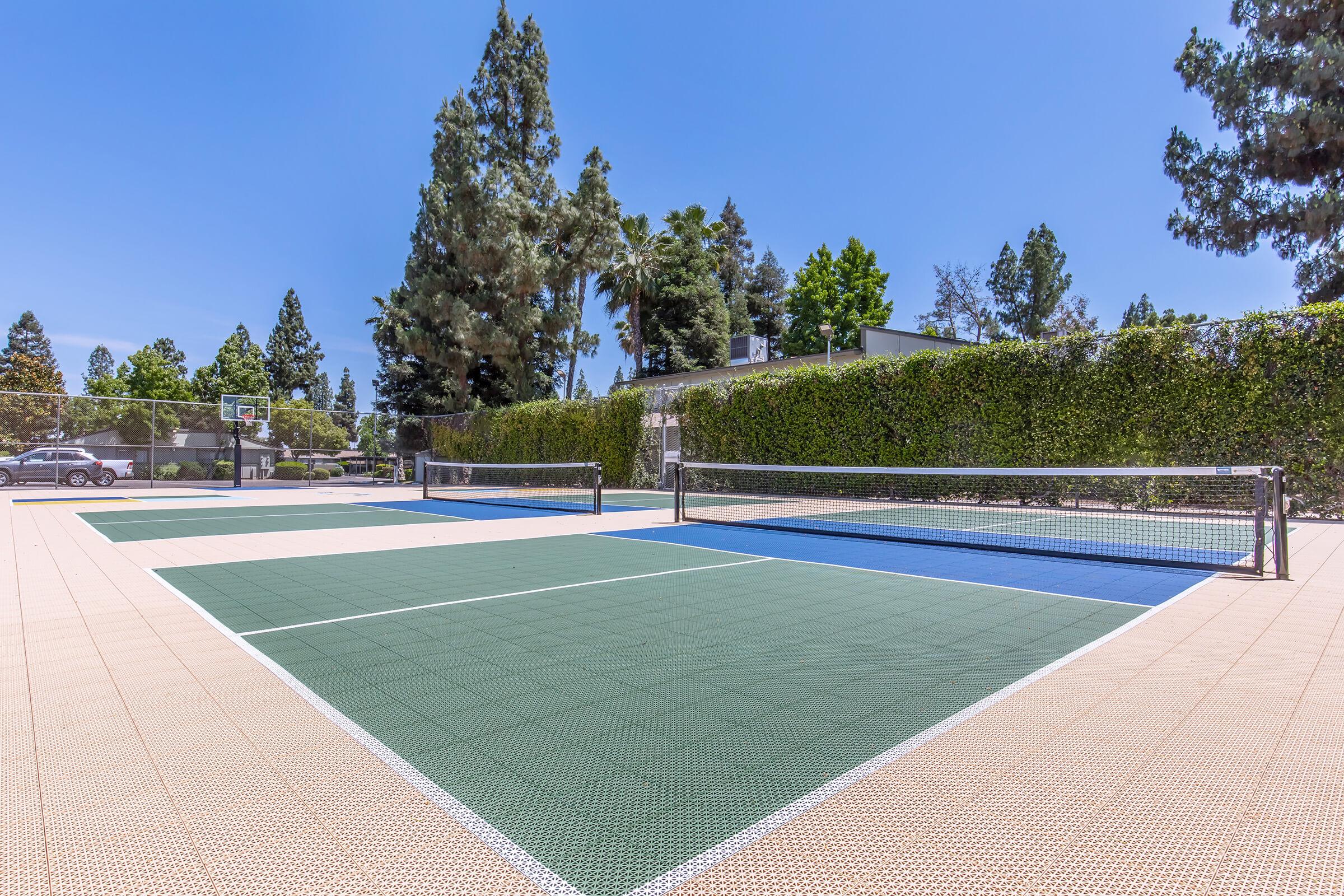 Two tennis courts with green and blue surfaces, surrounded by tall trees and hedges, under a clear blue sky. A basketball hoop is visible in the background, adding to the recreational setting. The area appears well-maintained and inviting for sports activities.