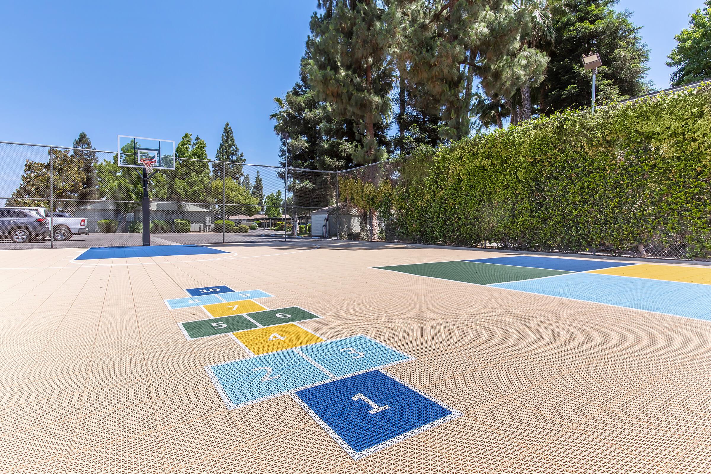A brightly lit outdoor play area featuring a hopscotch grid with numbered squares on a textured surface, adjacent to a basketball court with a hoop. Tall trees and greenery border the space, creating a vibrant and inviting atmosphere for recreational activities.