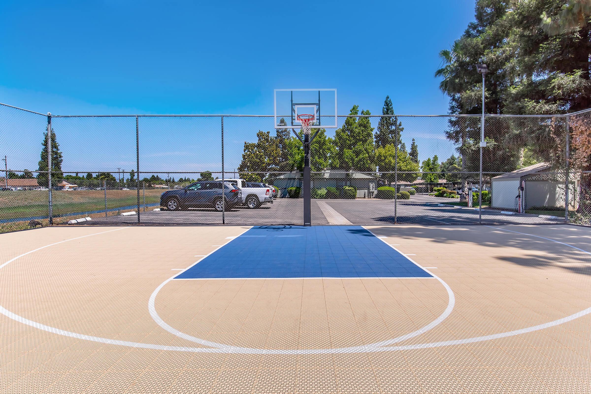 A basketball court with a blue section in the center, surrounded by a fence. In the background, there are trees and parked cars. The sky is clear and blue, indicating a sunny day.
