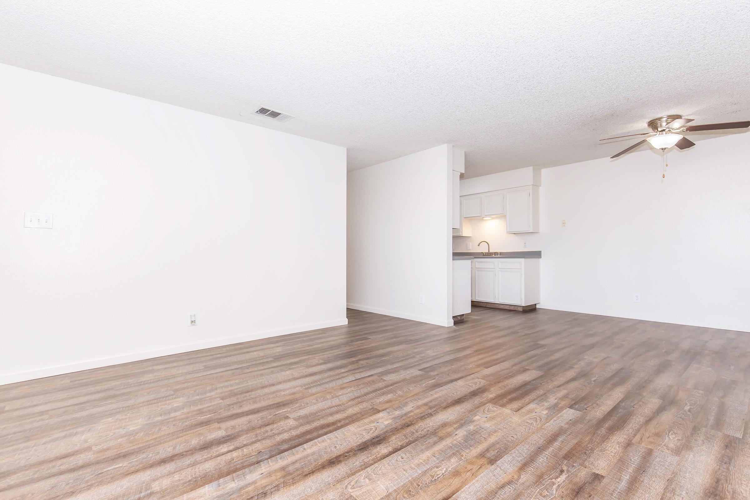 A spacious, empty living room featuring light-colored walls, a ceiling fan, and hardwood-style flooring. In the background, a kitchen area with white cabinetry and a counter is visible, creating an open layout. The room is well-lit and ready for furnishing.