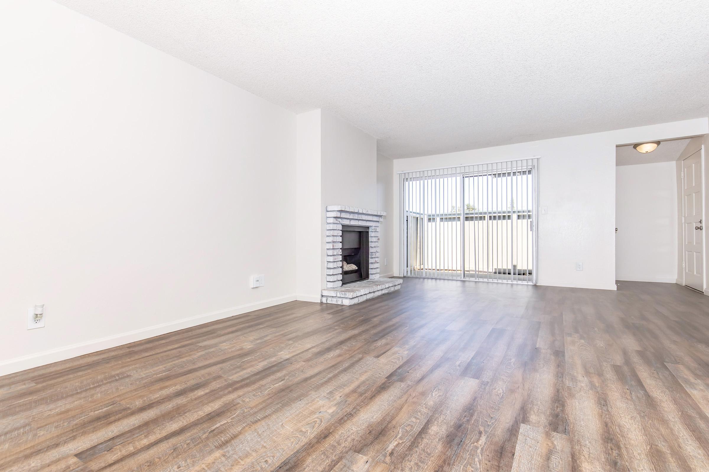 A spacious, empty living room with light-colored walls and a wooden floor. There's a white-brick fireplace on the left side and large sliding glass doors leading to a balcony or outdoor area. Natural light fills the room, and an adjacent doorway suggests access to another space.