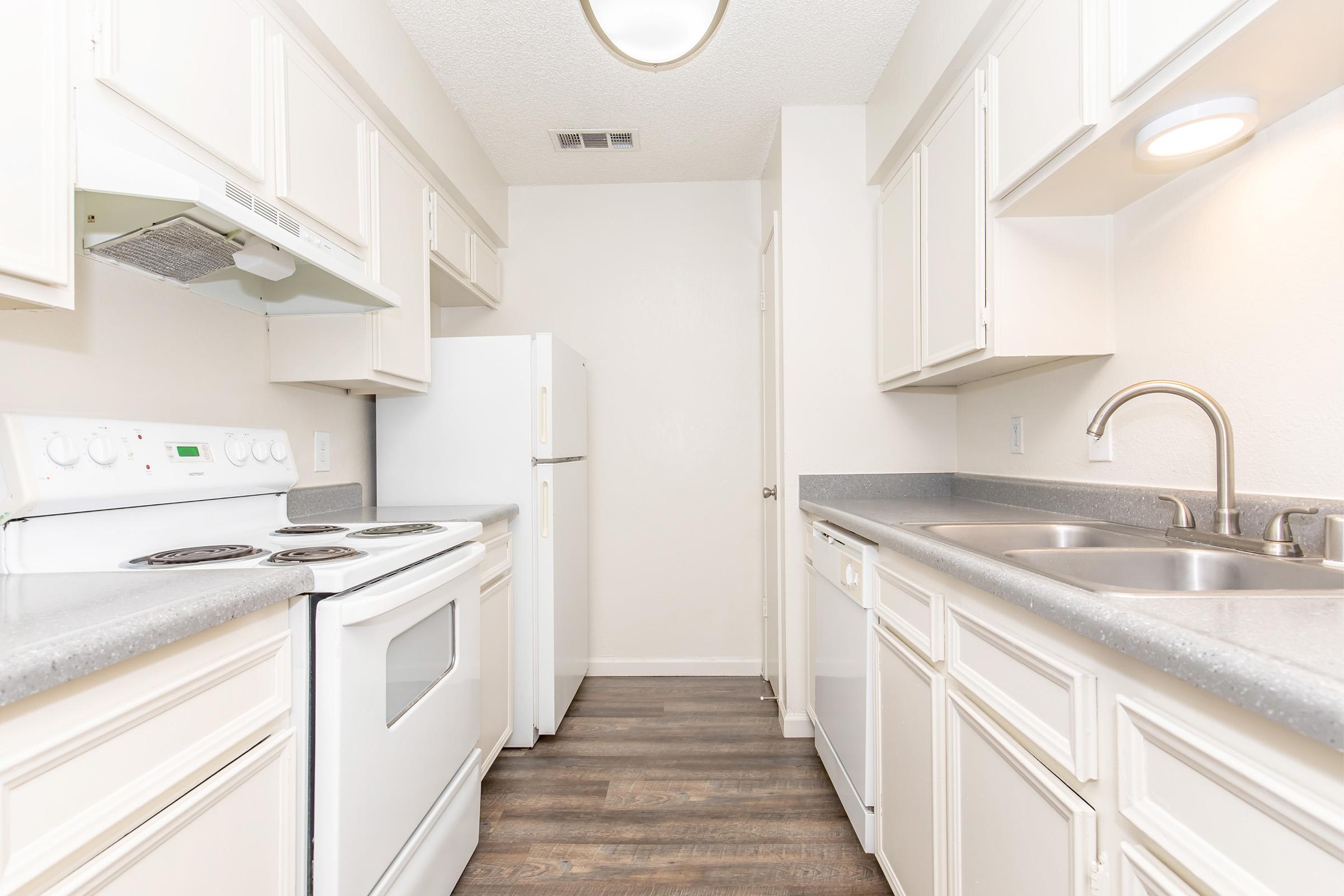 A bright, modern kitchen featuring white cabinetry and appliances, including a refrigerator, stove, and microwave. The countertops are gray, with a double sink and a dishwasher. The floor is a light wood finish, and the walls are a neutral tone, creating a clean and spacious atmosphere.
