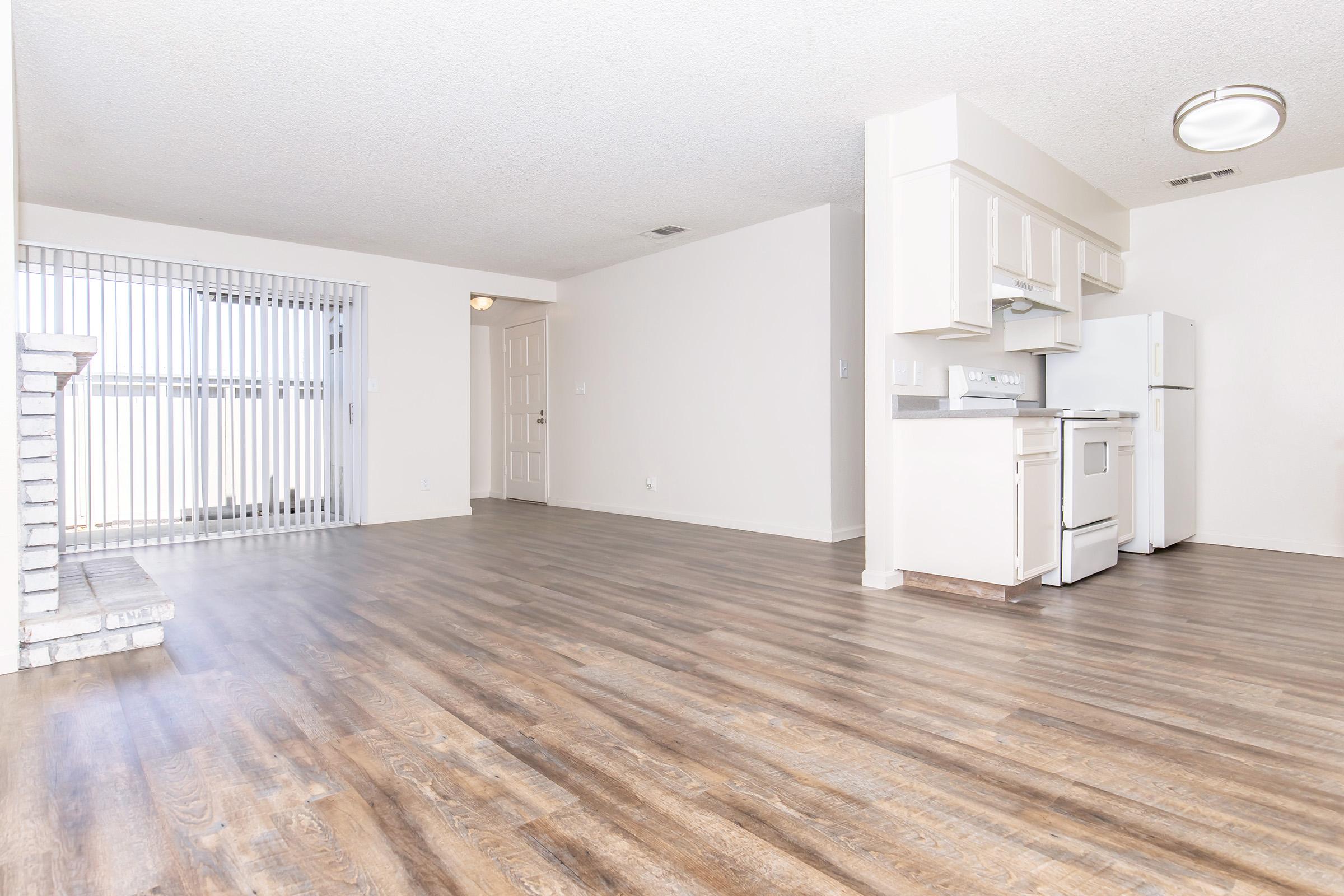 A spacious, empty living area featuring laminated wood flooring. There's a white kitchen with appliances visible, and a partially open layout connecting to a door leading outside. A barred window adds natural light to the space, which has neutral-colored walls and a ceiling light fixture.