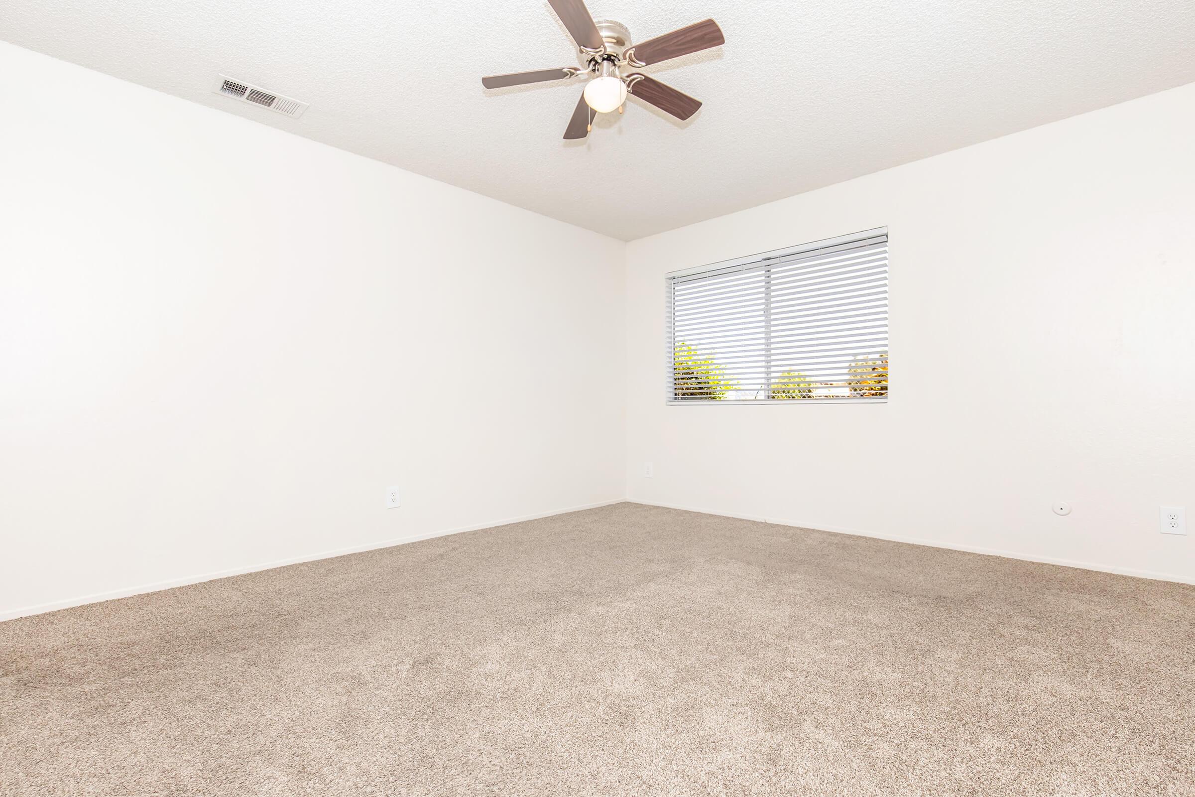 A spacious, empty room with light beige carpet, a ceiling fan, and a window covered by blinds. The walls are painted a light color, creating a bright and airy atmosphere. Natural light filters through the window, illuminating the room, which has no furniture or decorations.