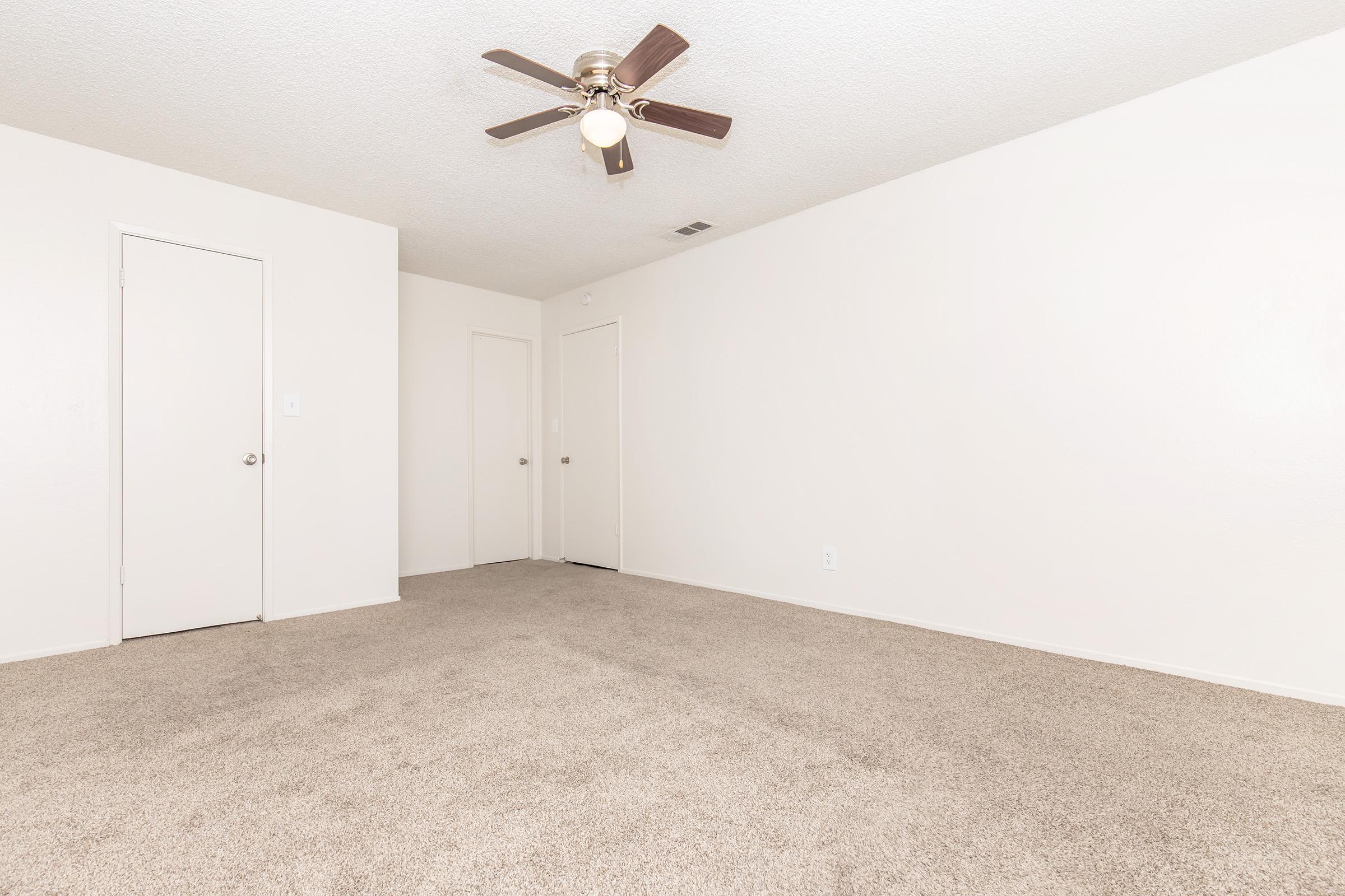 Spacious, empty room with light beige carpet and white walls. A ceiling fan with wooden blades is mounted in the center, and there are two closed doors on the left and right sides of the image, leading to other areas of the space. The room has a clean and simple aesthetic.