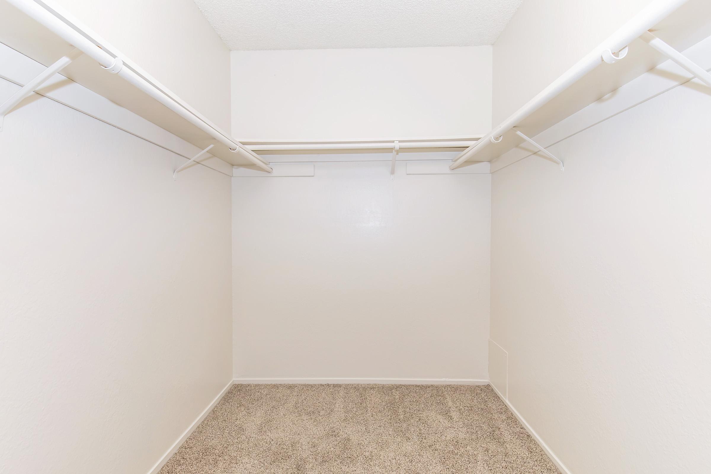 Empty walk-in closet featuring white walls and two horizontal clothing rods along the top. The floor is covered with carpet, and there are no shelves or additional storage items present. The overall appearance is clean and simple, with ample space for organizing clothes or other items.
