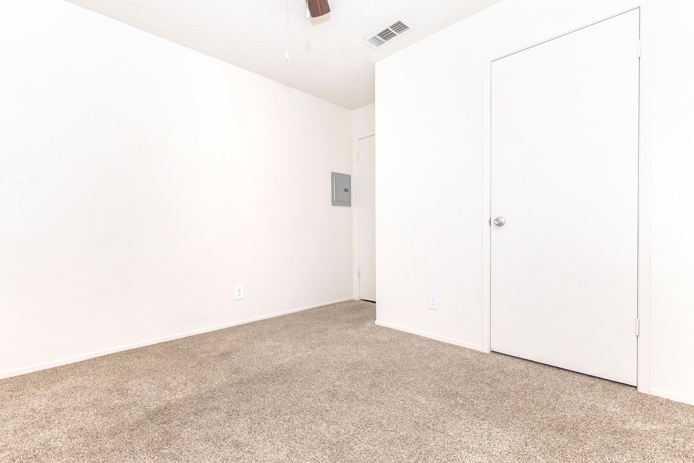 A spacious, empty room with light beige carpet and white walls. There is a closed door on the right and a small wall-mounted mailbox on the left. The ceiling has a fan fixture, and the overall ambiance is bright and simple, suggesting potential for personalization.