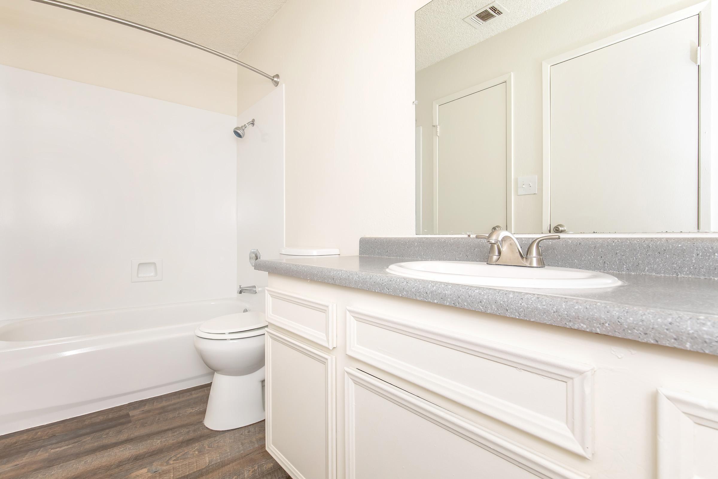A clean and simple bathroom featuring a white bathtub, toilet, and a gray countertop with a sink. The walls are light-colored, and the floor has a wood-like finish. There's a shower curtain rod installed, and a large mirror above the sink. Two doors are visible, likely leading to different rooms.