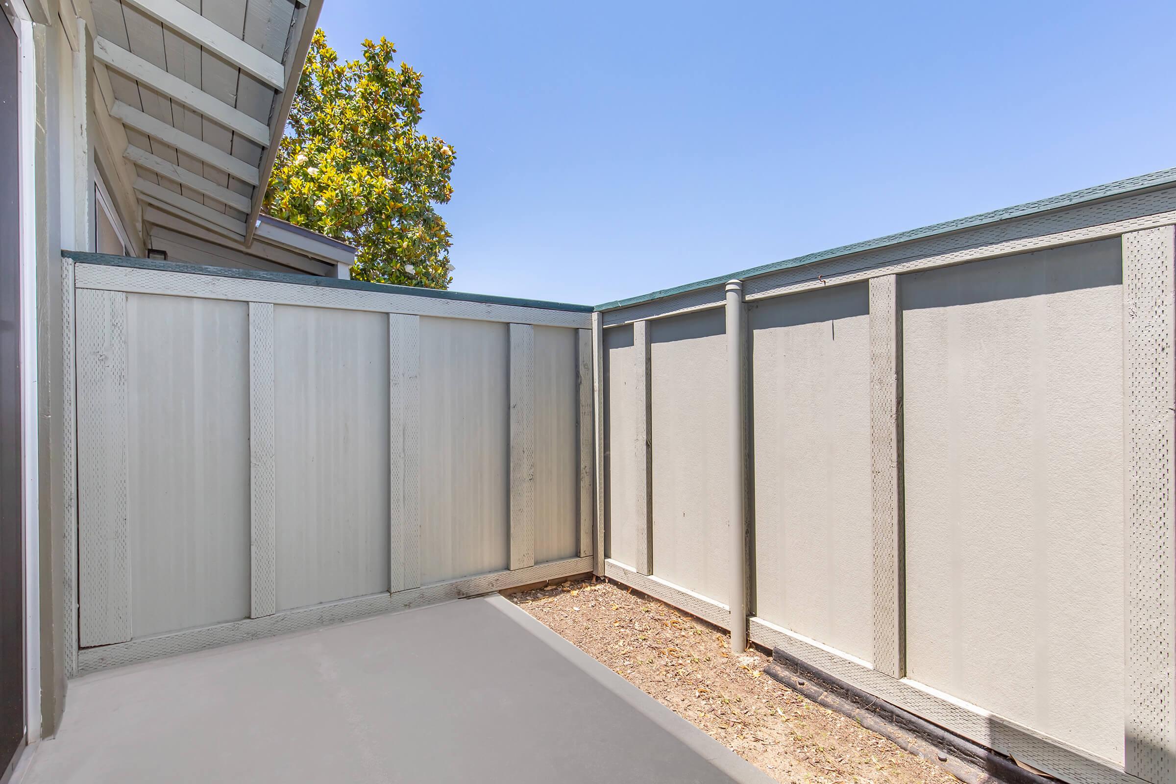 A fenced outdoor space with a gray concrete floor and wooden fencing. Bright blue sky visible above, with a tree partially seen in the background. The area appears clean and well-maintained, offering a private setting.
