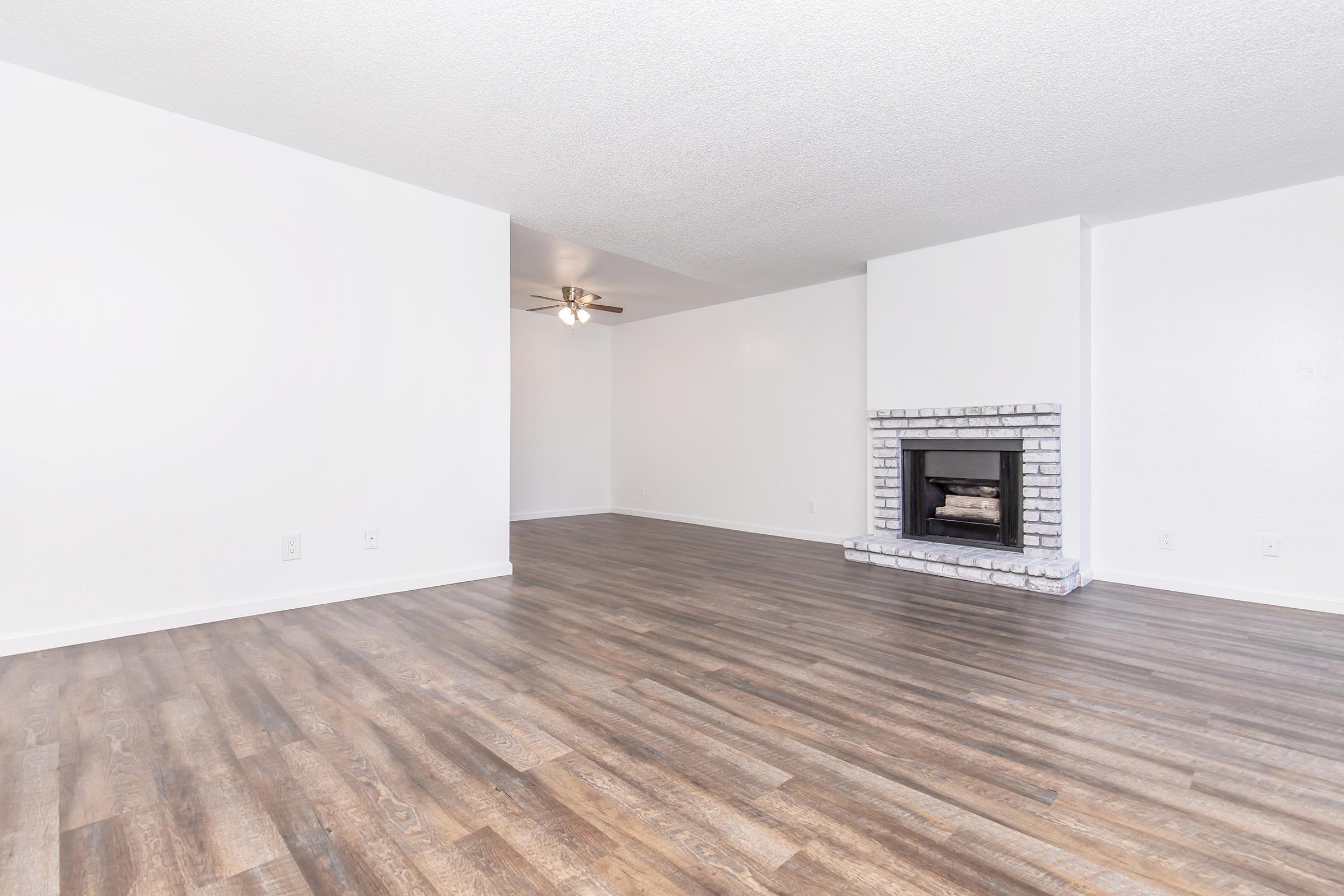 Empty interior room featuring light-colored walls, a ceiling fan, and a brick fireplace. The floor has a wood-like finish, and the space is well-lit, creating a bright and open atmosphere.