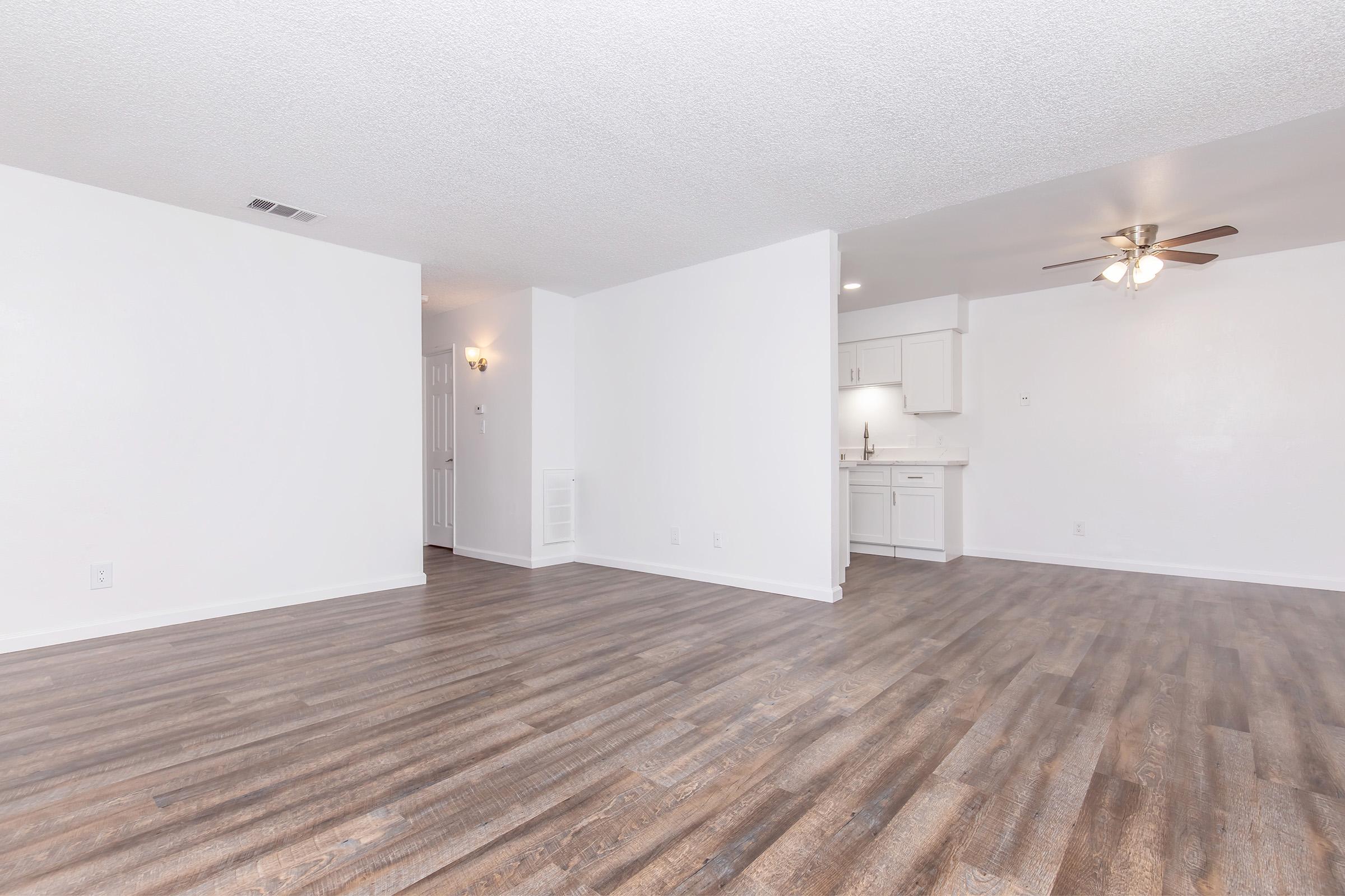 A spacious, empty living area with light-colored walls and a ceiling fan. Wood-like flooring extends throughout the room. In the background, a small kitchen area is visible with white cabinetry and a light countertop, contributing to a bright and airy atmosphere.