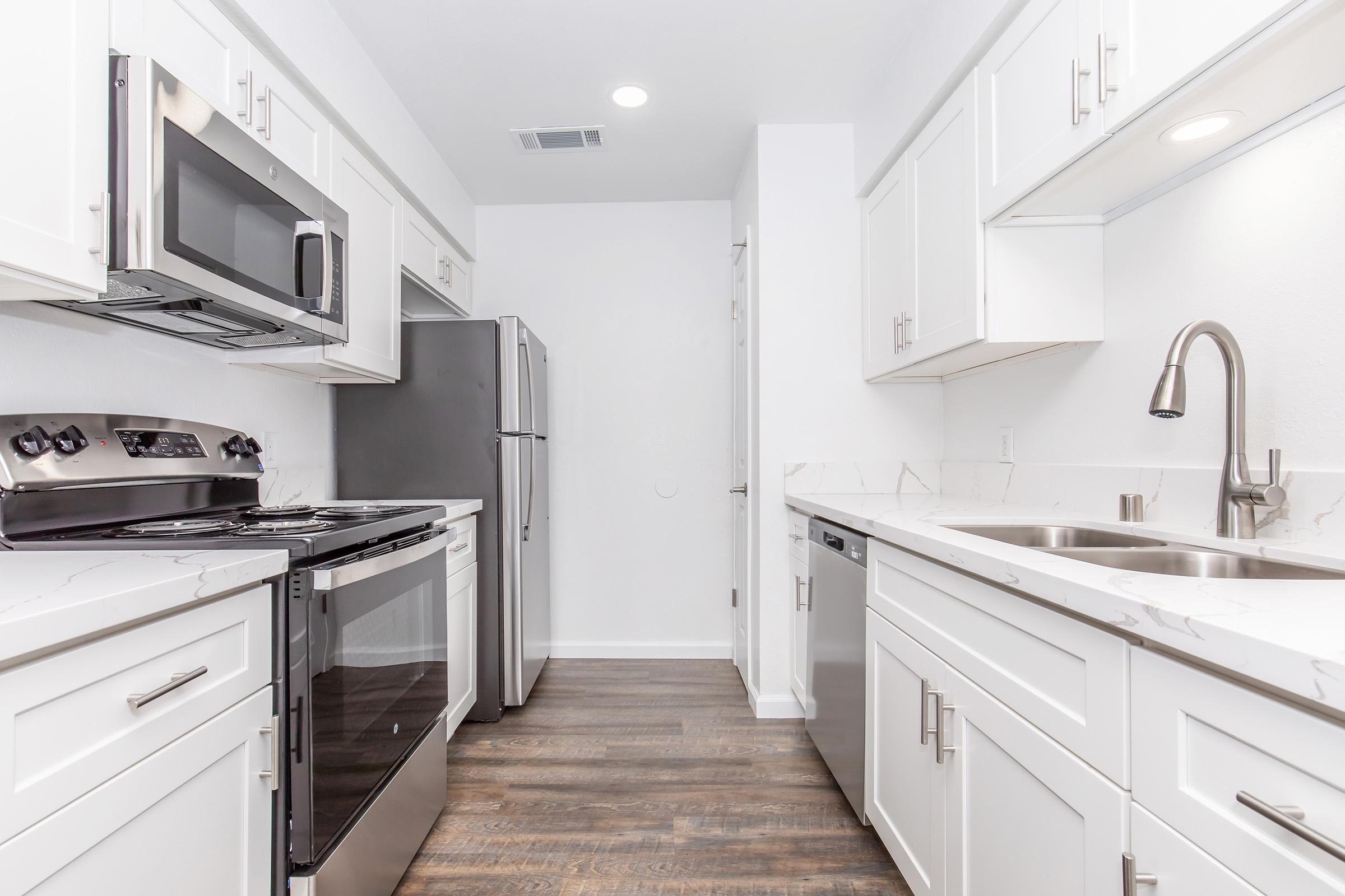 Modern kitchen featuring white cabinetry, stainless steel appliances including a microwave, stove, and refrigerator. The countertops are light-colored and have a marble-like finish. A double sink is located beside the dishwasher. The flooring is dark wood, providing a contrast to the bright, clean look of the kitchen.