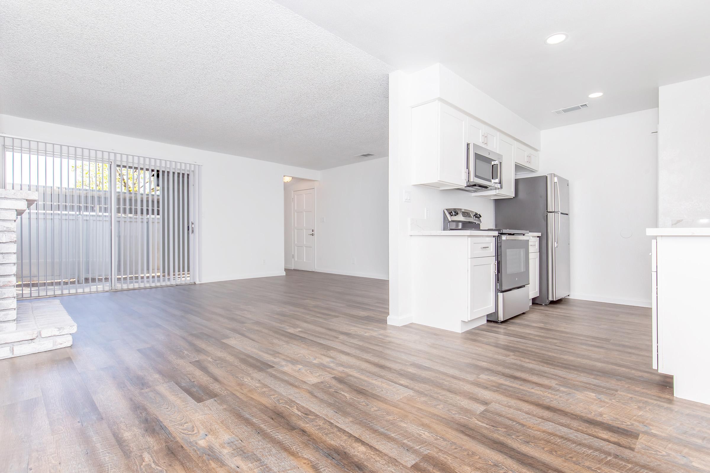 A bright, open-concept living space featuring a kitchen with stainless steel appliances and a dining area. The floor is finished with wood-like laminate, and there are large windows with vertical blinds allowing natural light. A sliding glass door leads to an outdoor area. The walls are painted white, creating a fresh, modern atmosphere.