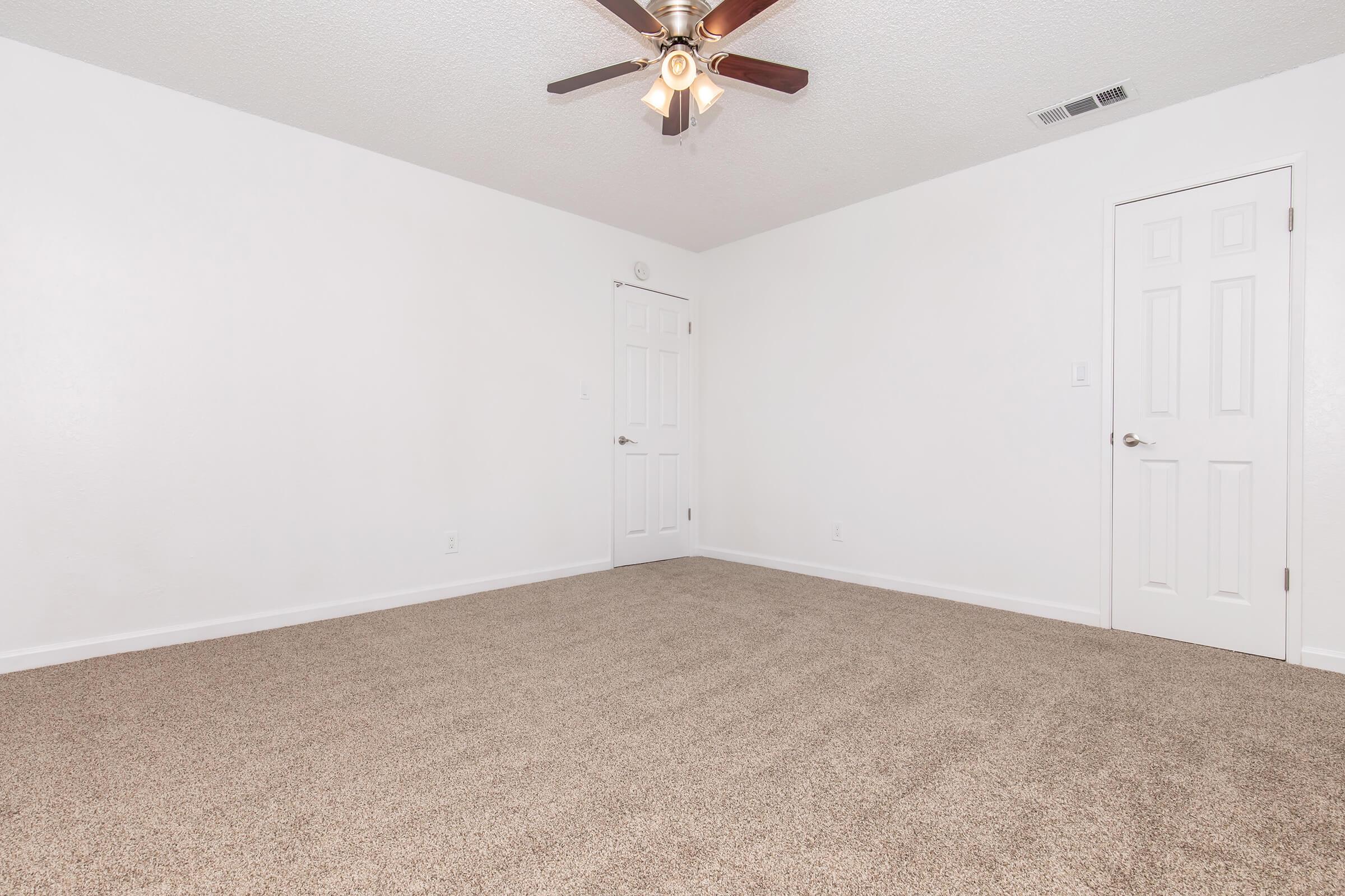 A spacious, empty room with light beige carpet and white walls. It features a ceiling fan with wooden blades and a single light fixture. Two doors are visible: one leading to a closet and the other to another part of the space. The room is well-lit, creating a bright and airy atmosphere.