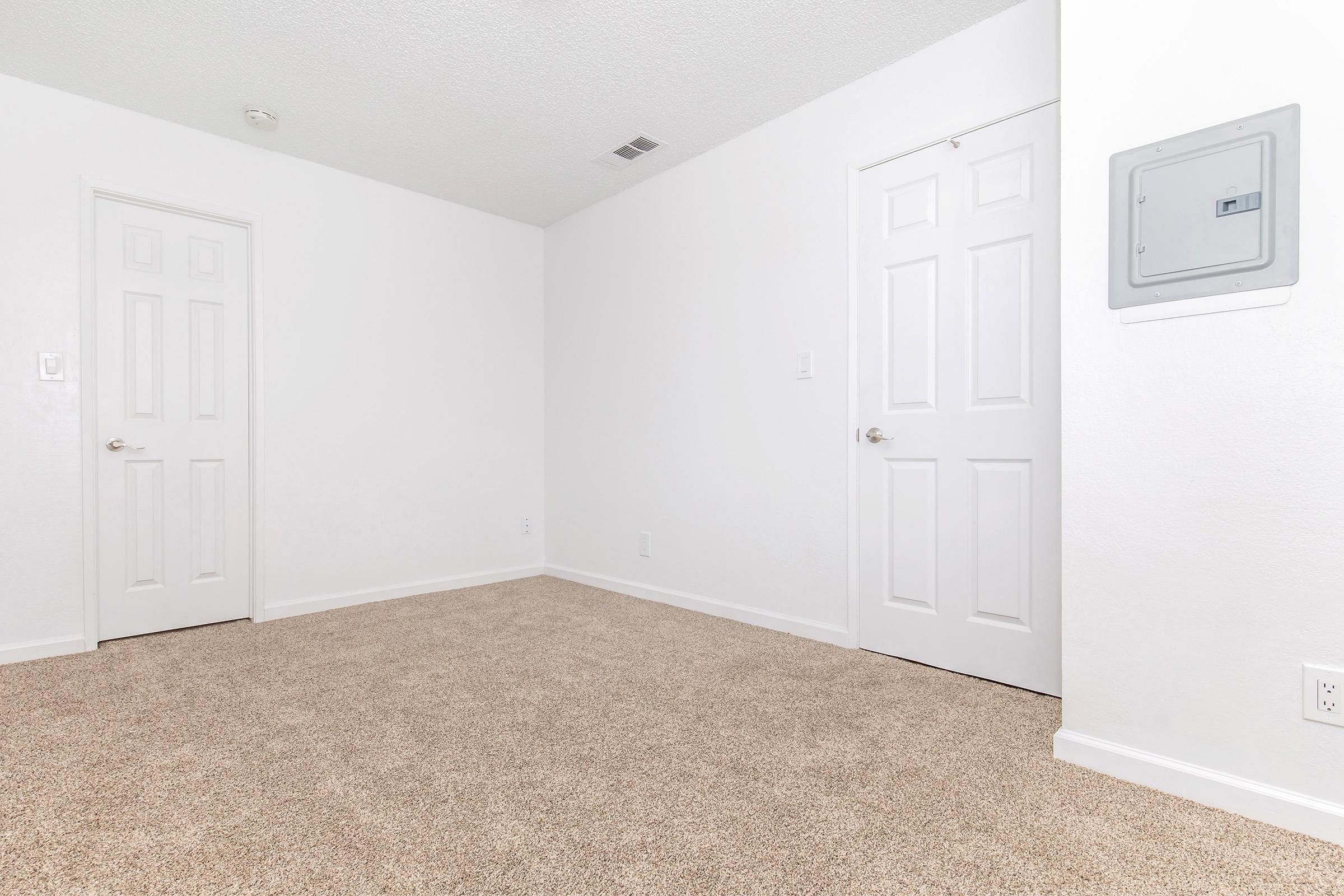 A spacious, empty room featuring light beige carpet flooring, white walls, and two closed doors on the left. A wall-mounted electrical panel is visible on the right side, and the overall atmosphere is bright and uncluttered, suggesting a fresh, inviting space ready for furnishing.