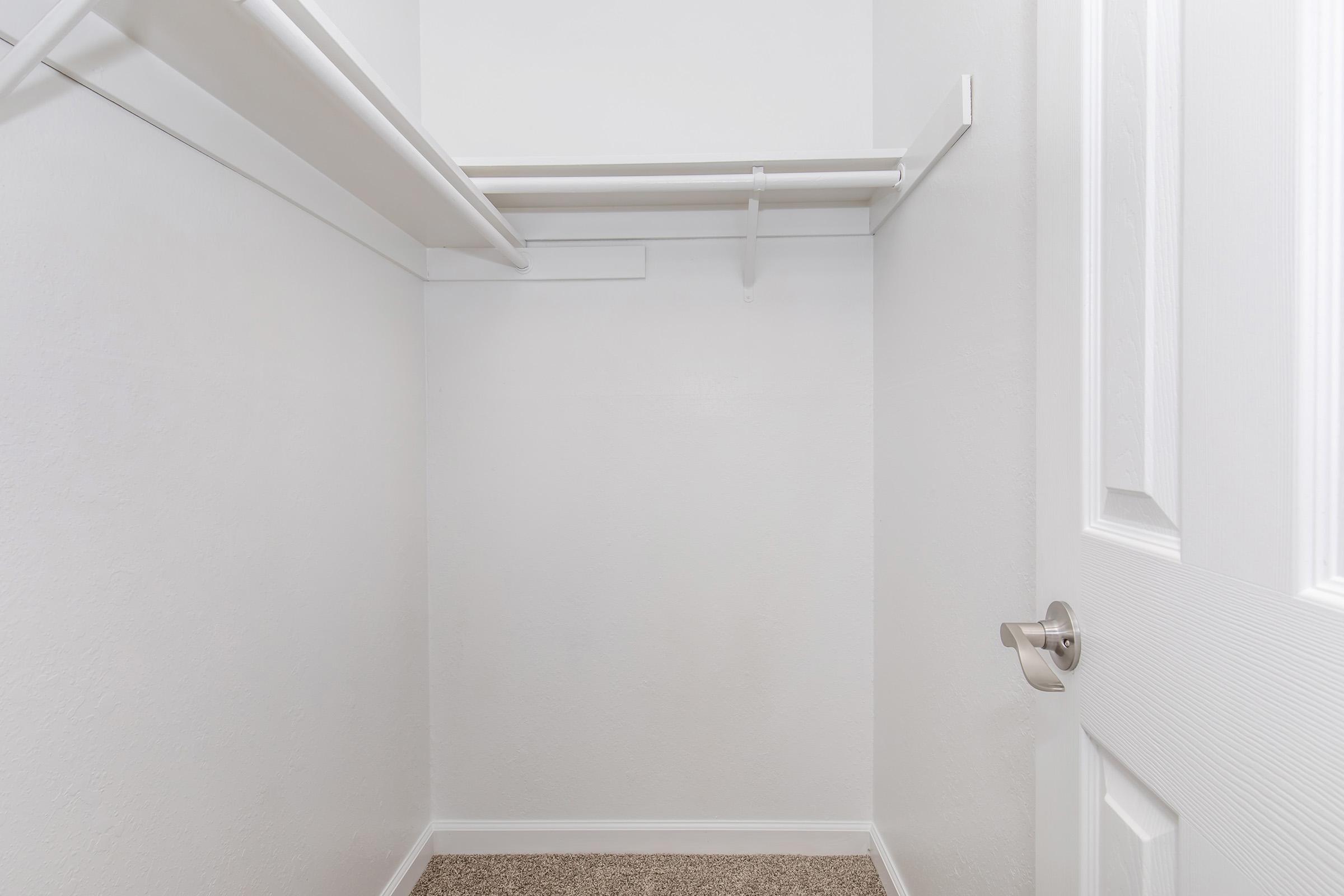 Empty closet with white walls and a light carpeted floor. A single shelf is mounted on the wall, and a hanging rod is visible. The closet door is partially closed, revealing the interior and emphasizing its spaciousness. The overall appearance is clean and uncluttered.