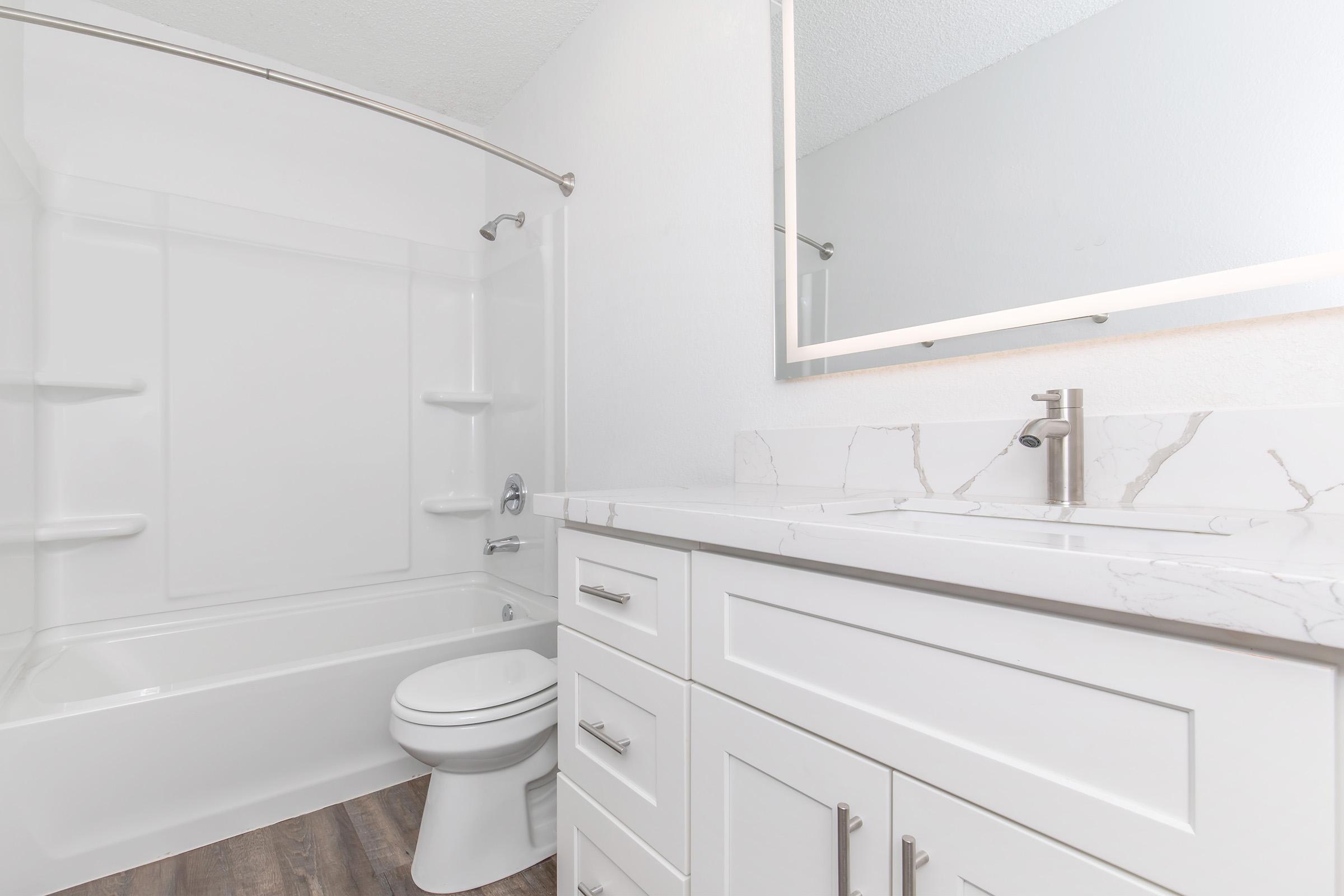 A clean and modern bathroom featuring a white tub and shower combination, a sleek vanity with a marble countertop, and a mirror. The flooring is a dark wood laminate, and the walls are painted in a light color, creating a bright and spacious atmosphere.