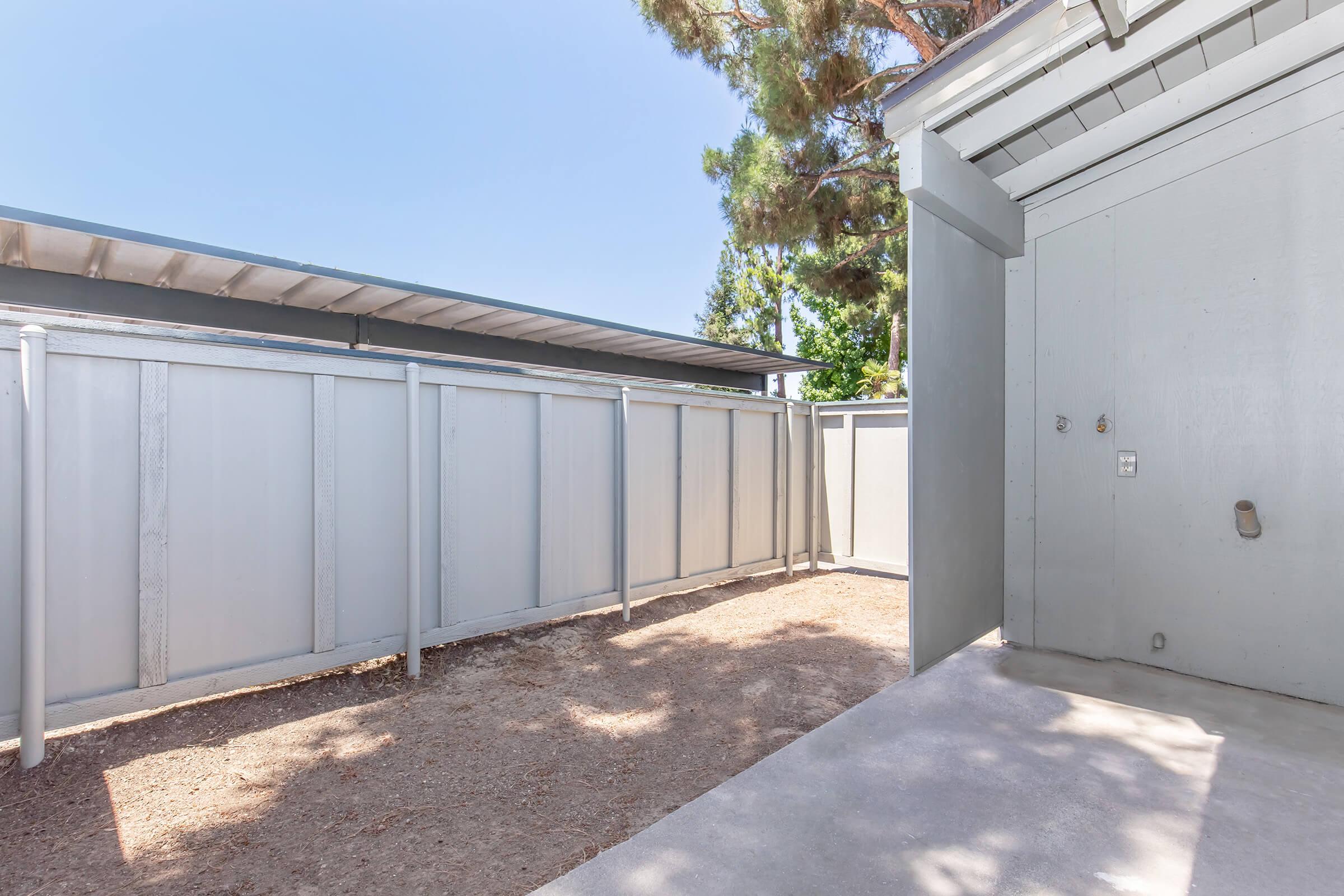 A fenced outdoor area featuring a gravel surface and a concrete pathway. There are two gray walls on either side, with one wall having a small door. The scene is well-lit, with trees visible in the background and clear blue sky overhead.