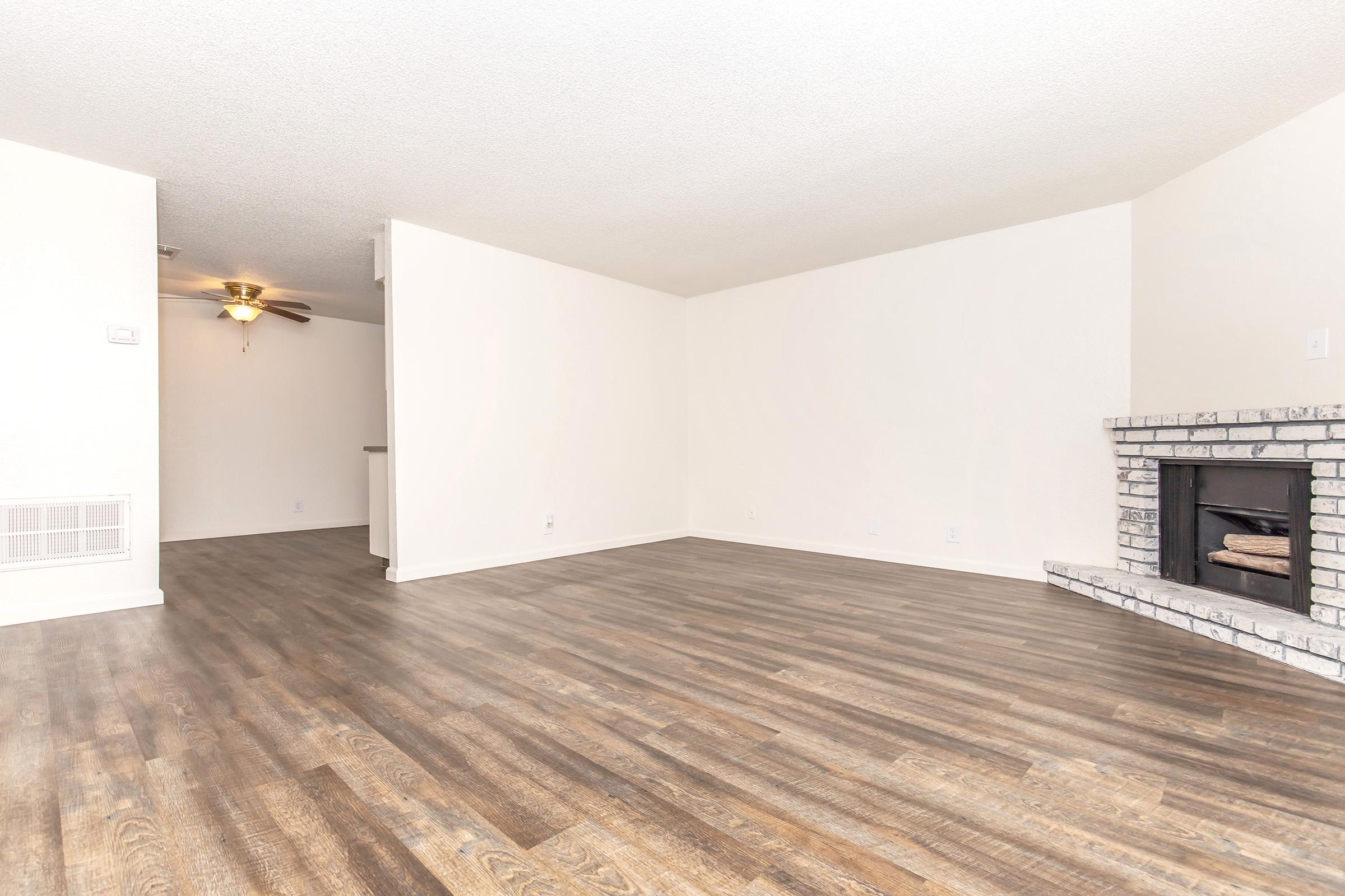 A spacious living room with light-colored walls and a wooden floor. There is a brick fireplace on the right side, and a ceiling fan is visible on the left. The room is empty, creating an open and airy atmosphere, with natural light flooding in.