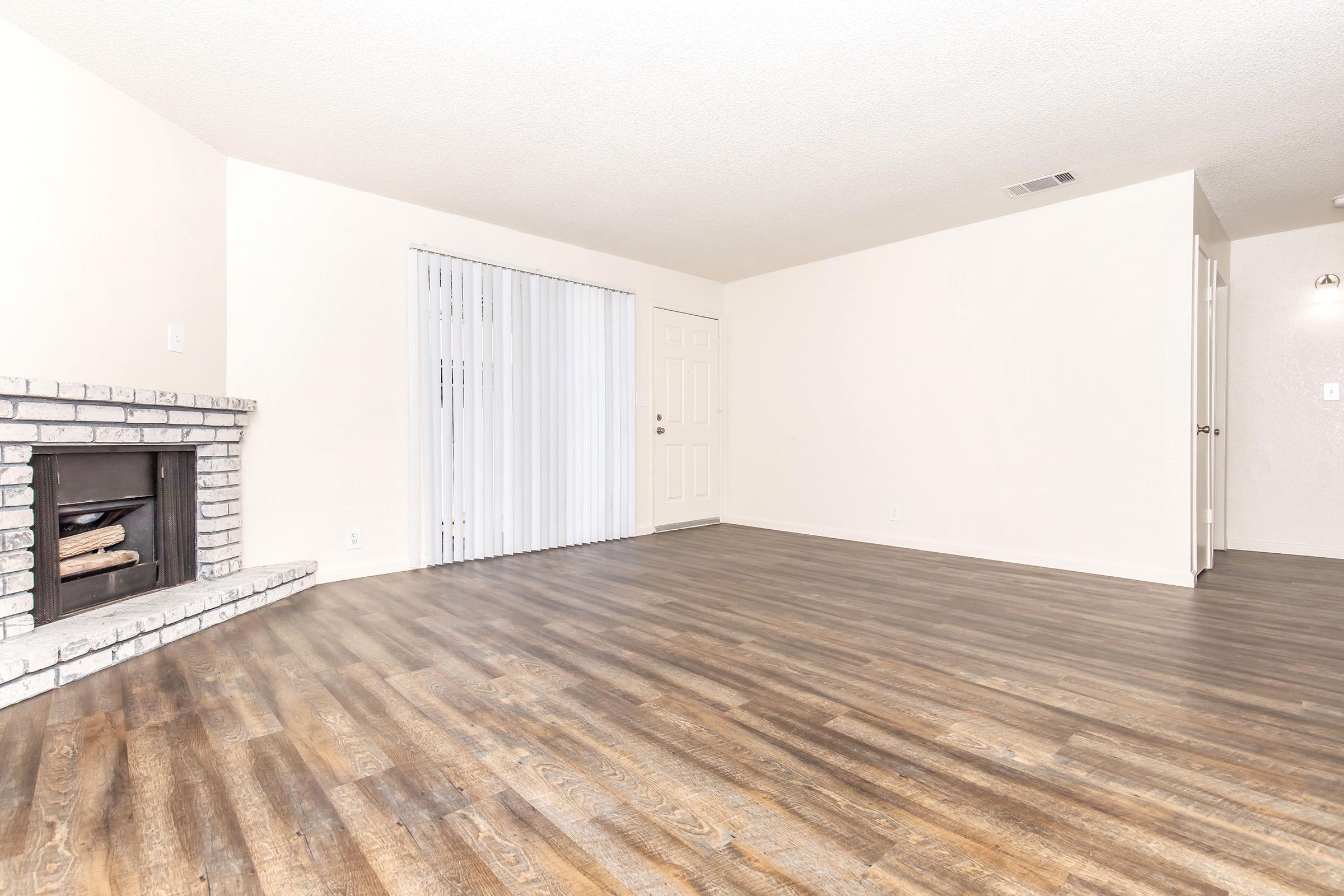 A spacious, empty living room featuring a light-colored wall, a brick fireplace, and sliding glass doors with vertical blinds. The floor has a wood-like finish, and there's a closed door leading to another room on the right. The overall ambiance is bright and inviting, with plenty of natural light.