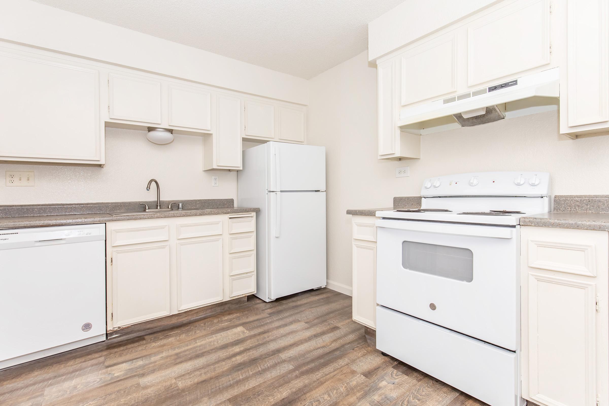 A clean, modern kitchen featuring white cabinetry, a refrigerator, a stove, and a dishwasher. The countertops are gray, and the floor has a wood-like appearance. The walls are painted a neutral color, creating a bright and inviting space.